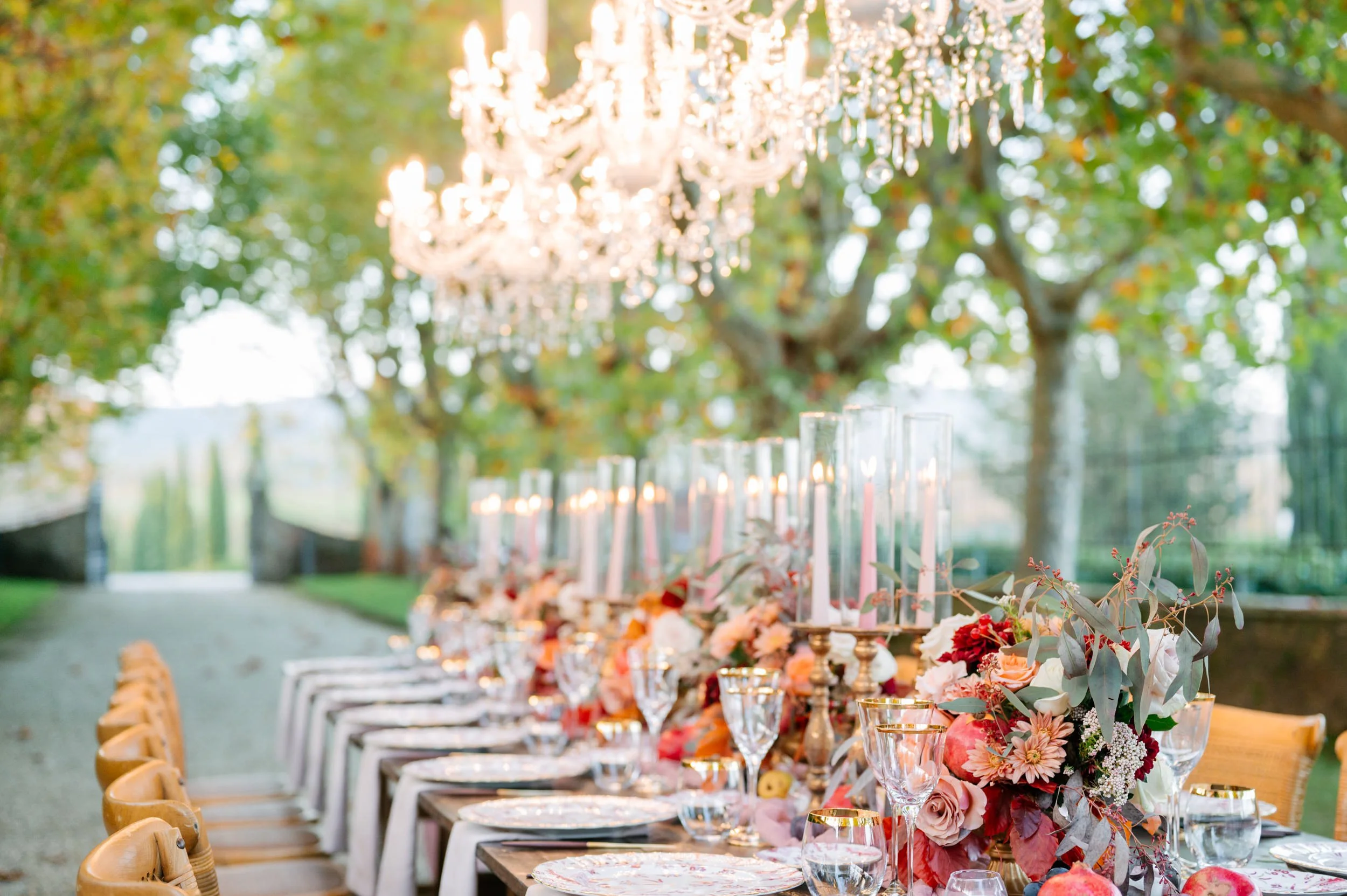 A long outdoor dining table decorated with floral arrangements, candles, and elegant tableware, set in a garden with trees and chandeliers overhead.