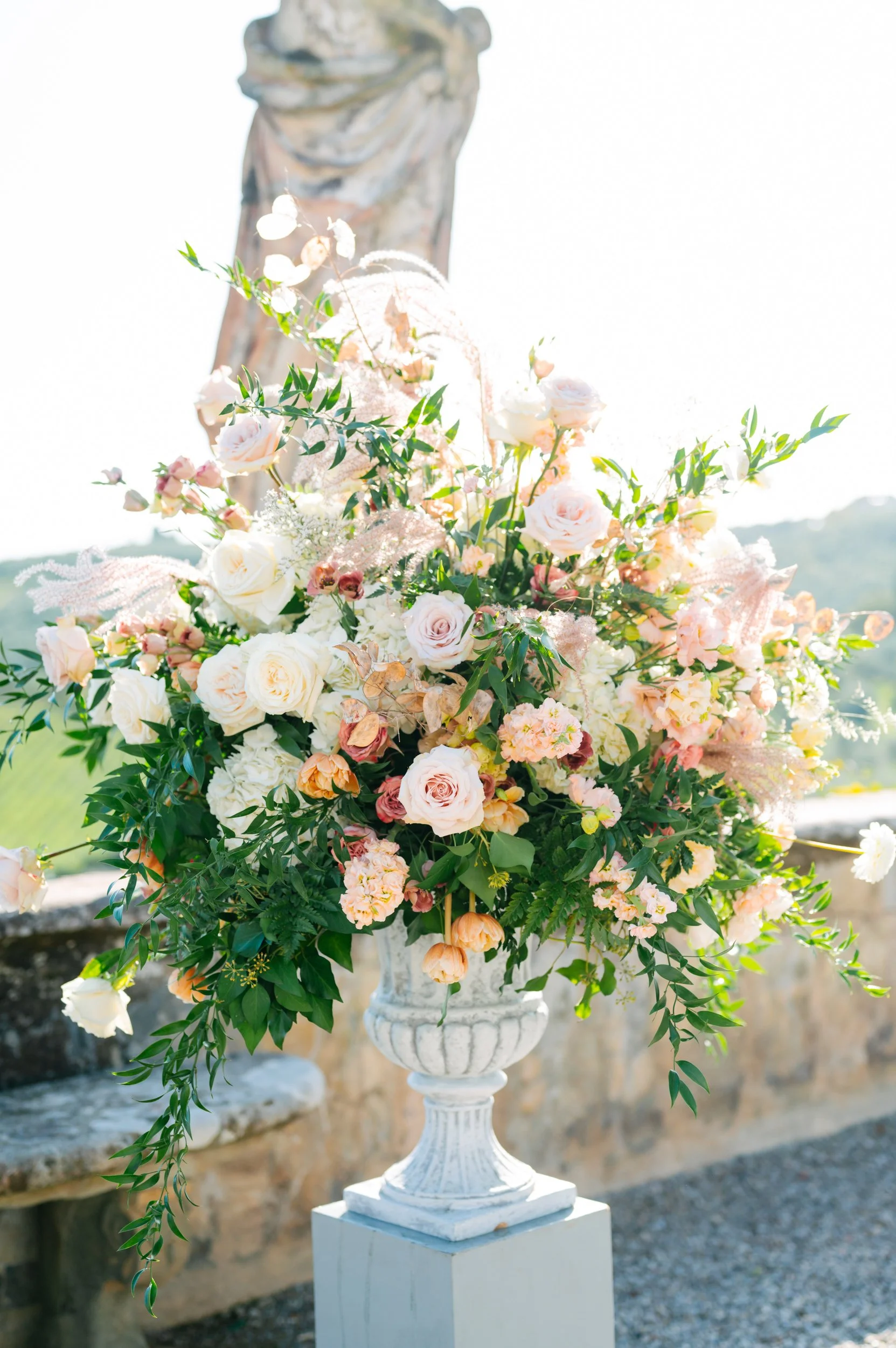 A large floral arrangement of pink and white roses, peach and pink flowers, and green foliage in a white urn-shaped vase outdoors with a stone wall and scenic background.