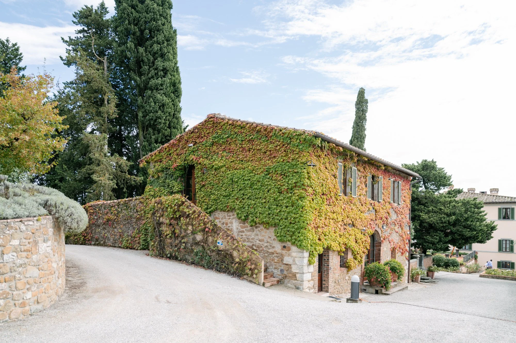 A rustic house covered in green and red ivy, situated on a curved gravel driveway with a stone wall and trees in the background under a partly cloudy sky. Luxury editorial wedding photography in Tuscany. Destination wedding in Italy. L&V Photography