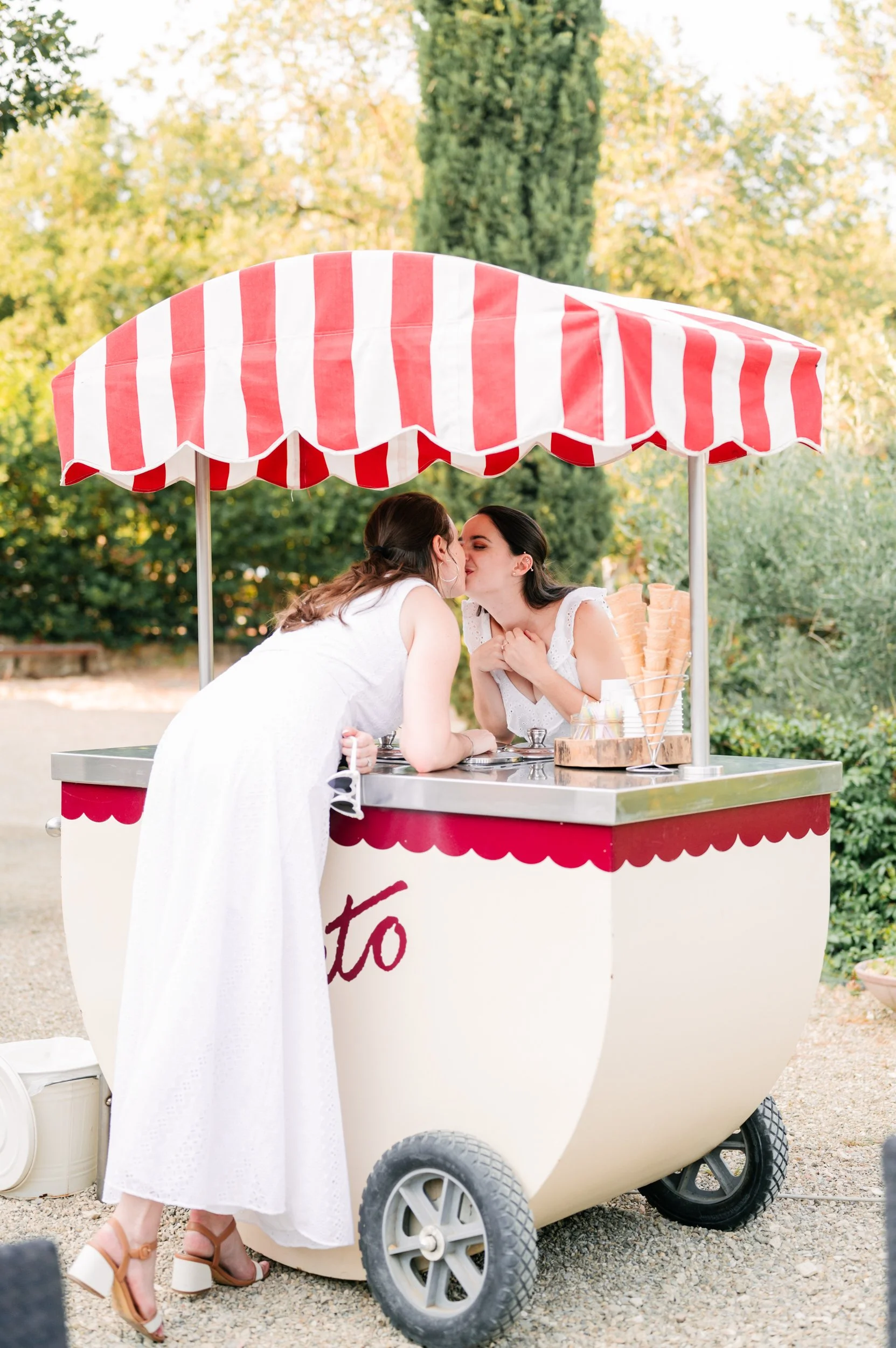 Two women at an ice cream cart sharing a kiss outdoors on a sunny day, surrounded by green trees.