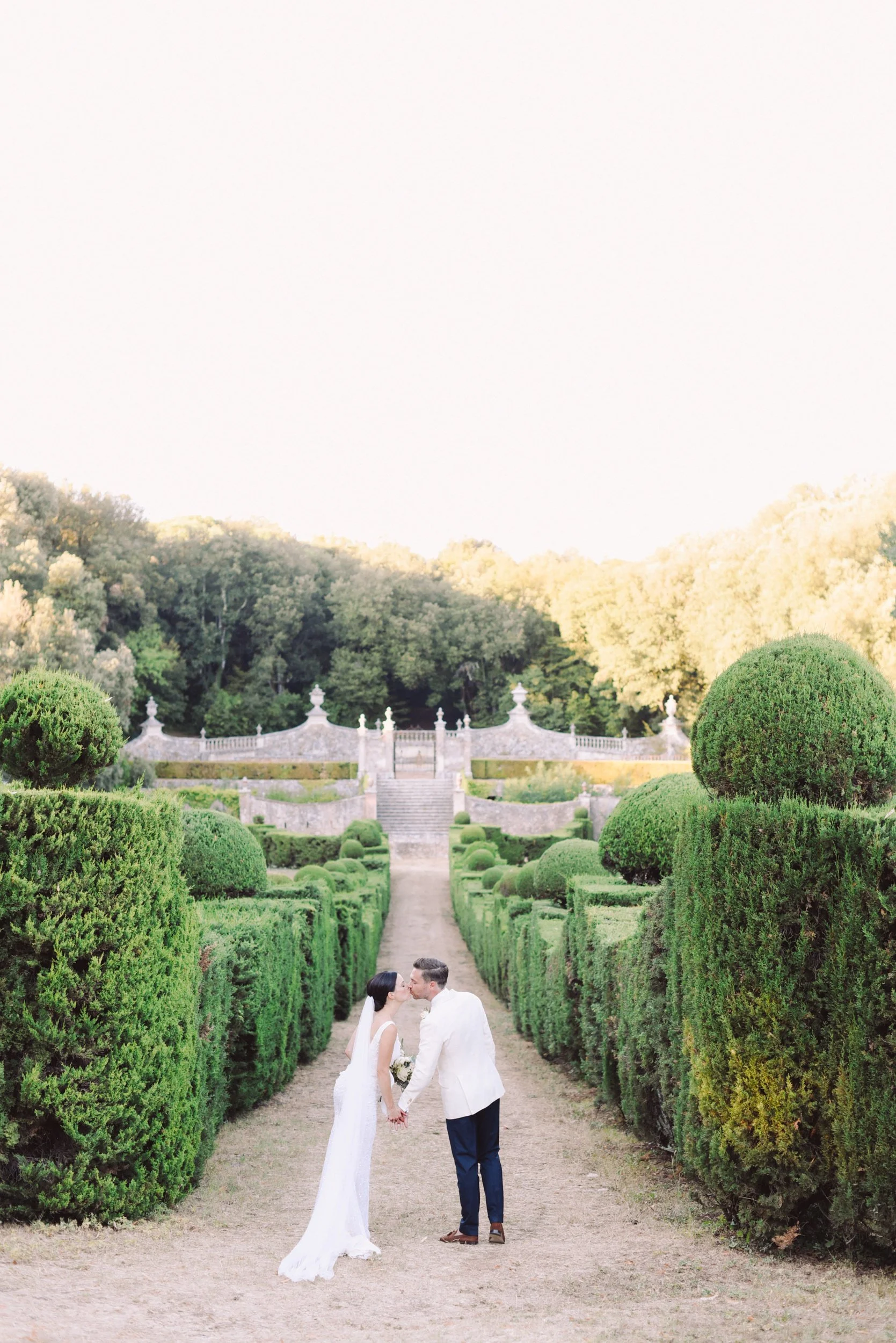 A bride and groom holding hands and kissing in a manicured garden with hedges and stairs.