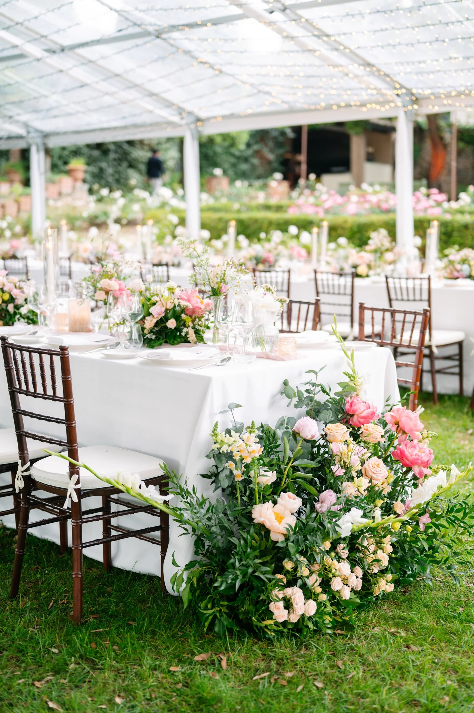 Decorated outdoor wedding reception with table settings, floral centerpieces, and string lights overhead.