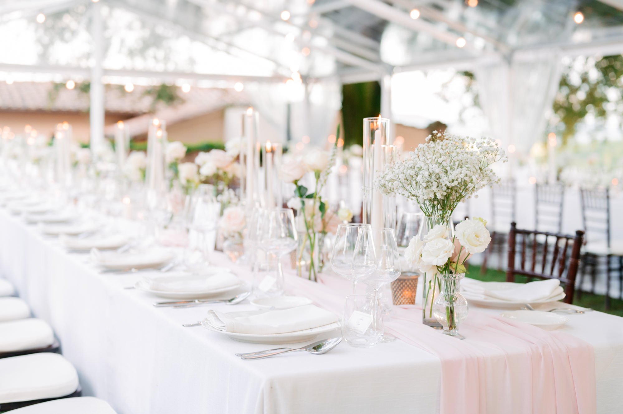 Elegant outdoor banquet table decorated with white flowers, candles, and glassware under a glass canopy.