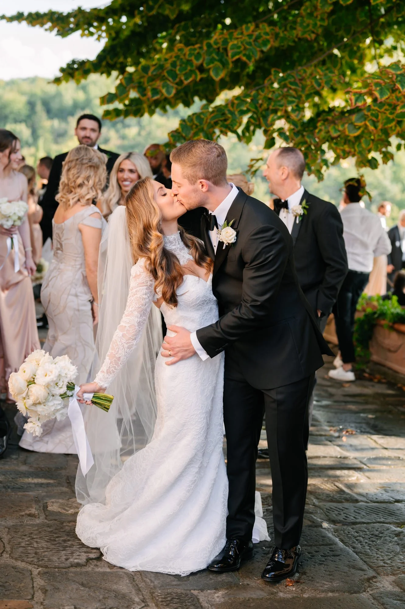 Bride and groom kissing during their wedding ceremony, surrounded by wedding guests and outdoor greenery.
