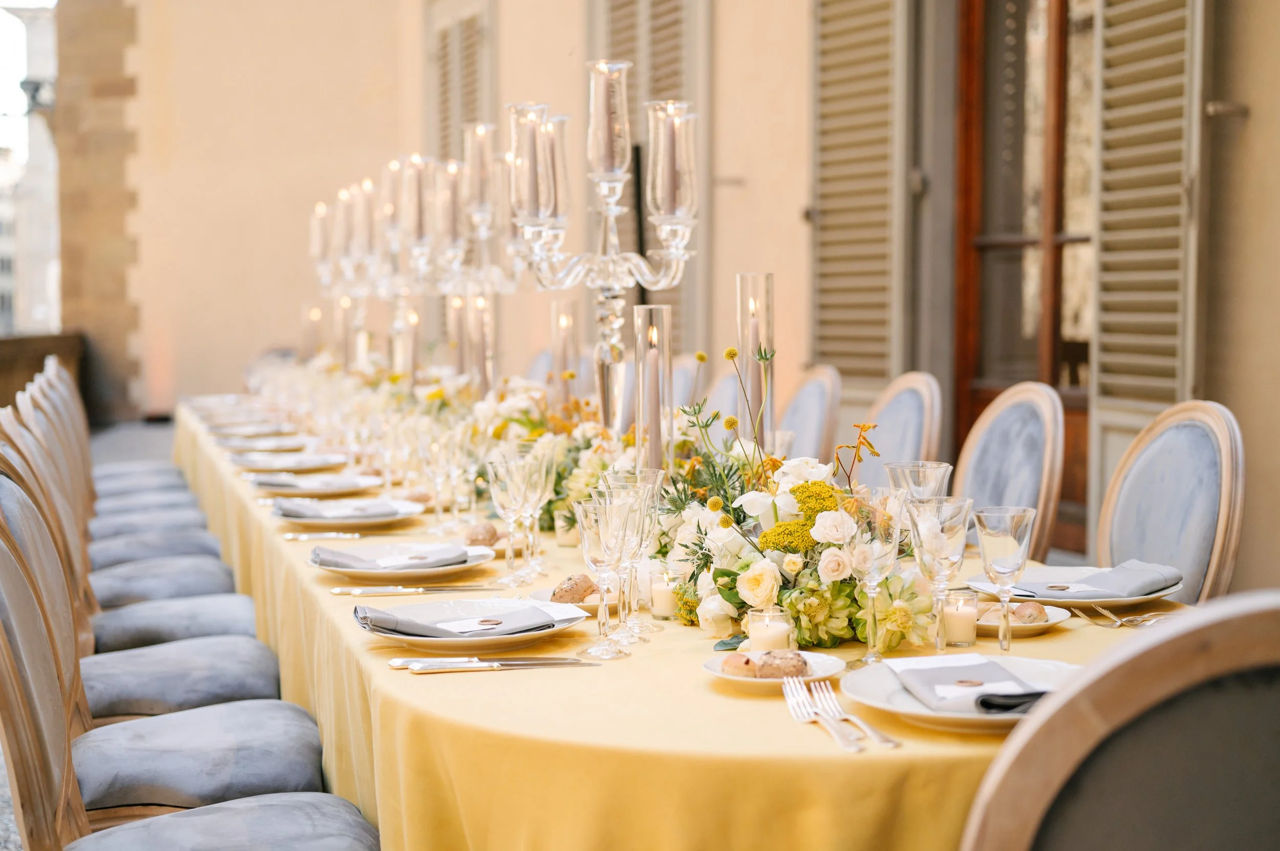 Elegant banquet table set with white plates, glassware, napkins, and a yellow tablecloth. The table is decorated with a floral centerpiece of white, yellow, and green flowers and tall glass candelabras.