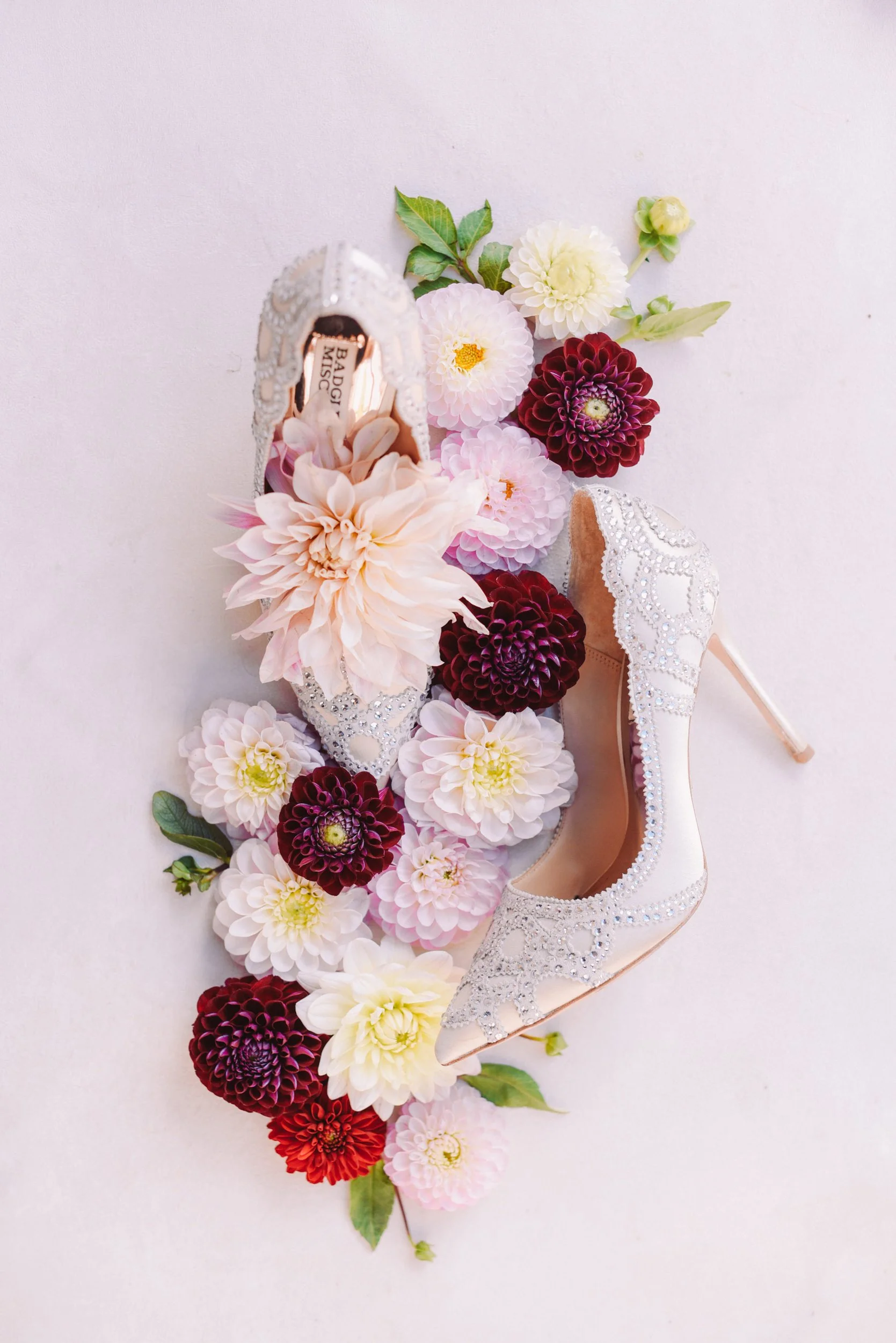 A pair of embellished high-heeled shoes surrounded by pink, red, purple, and white flowers, all arranged on a white surface.