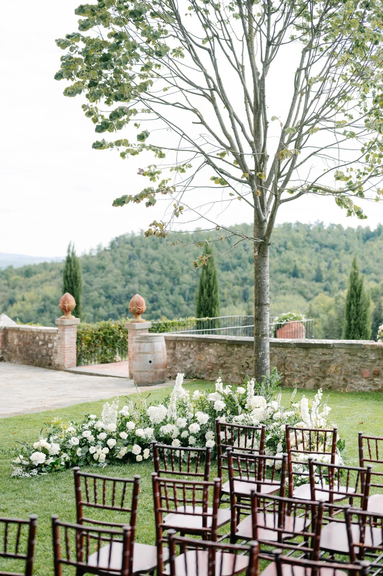 Outdoor wedding setup with white floral arrangements under a tree, wooden chairs arranged for guests, stone wall with decorative urns, and scenic hillside view in the background.