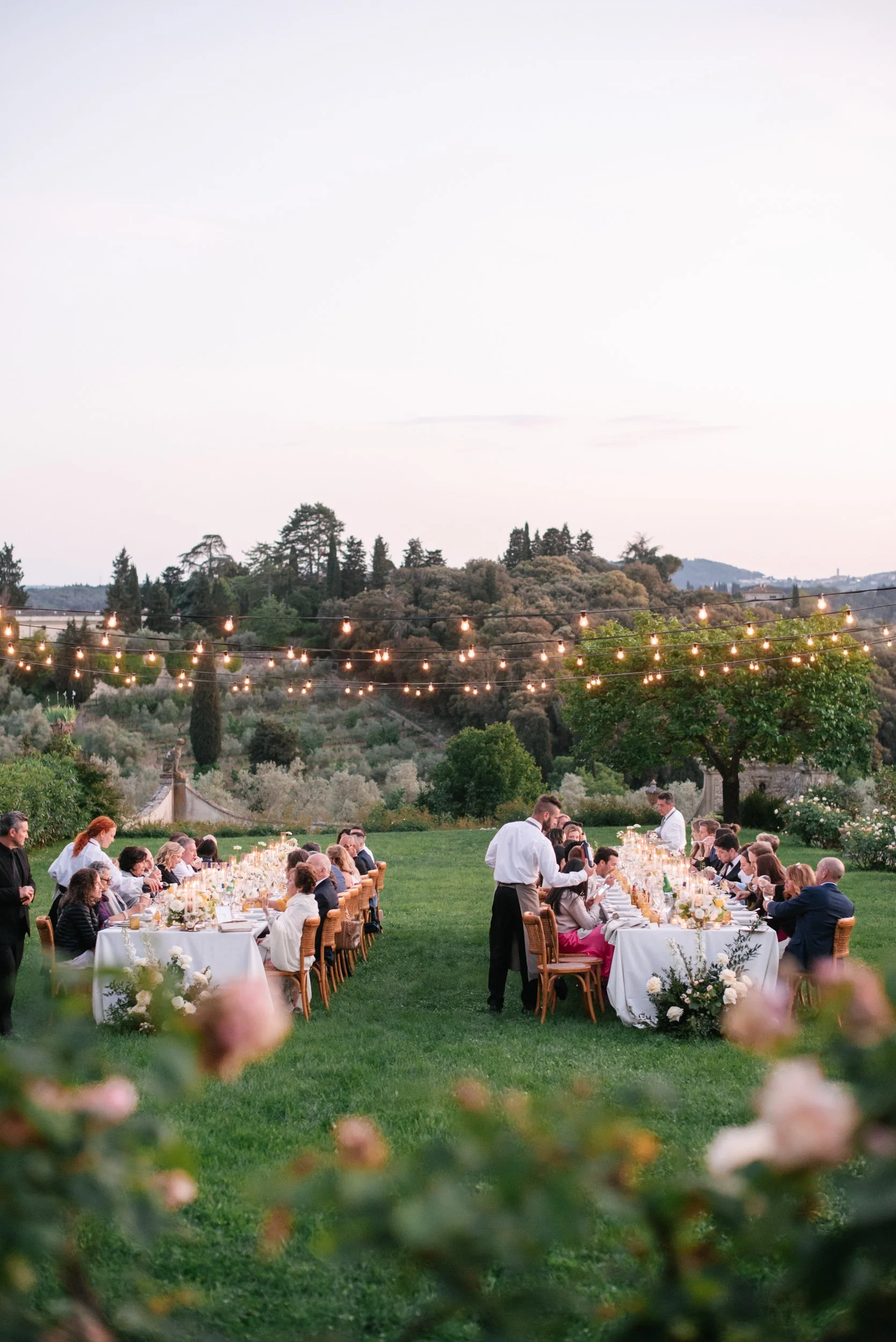 Outdoor wedding reception at sunset with long tables, guests, string lights, greenery, and scenic hills in the background.