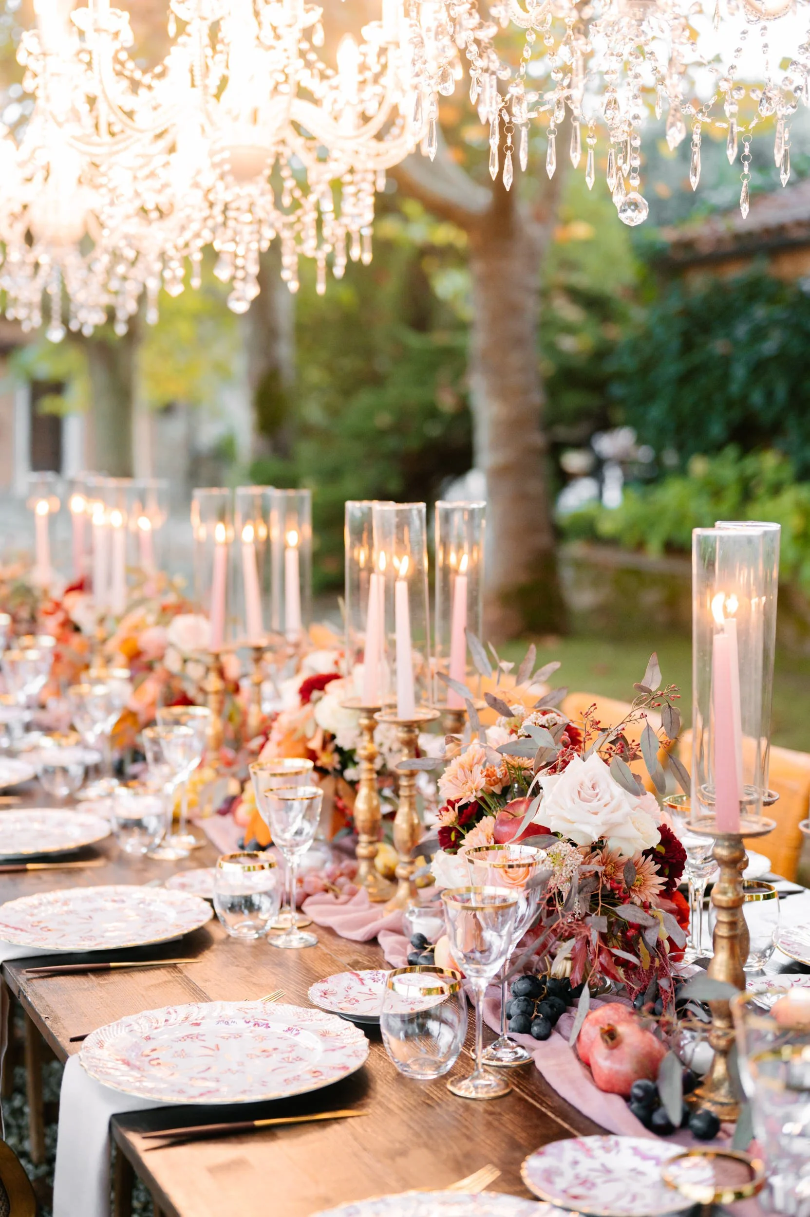 An elegant outdoor wedding reception table decorated with floral arrangements, candles in glass holders, and a hanging crystal chandelier, set in a garden during sunset.