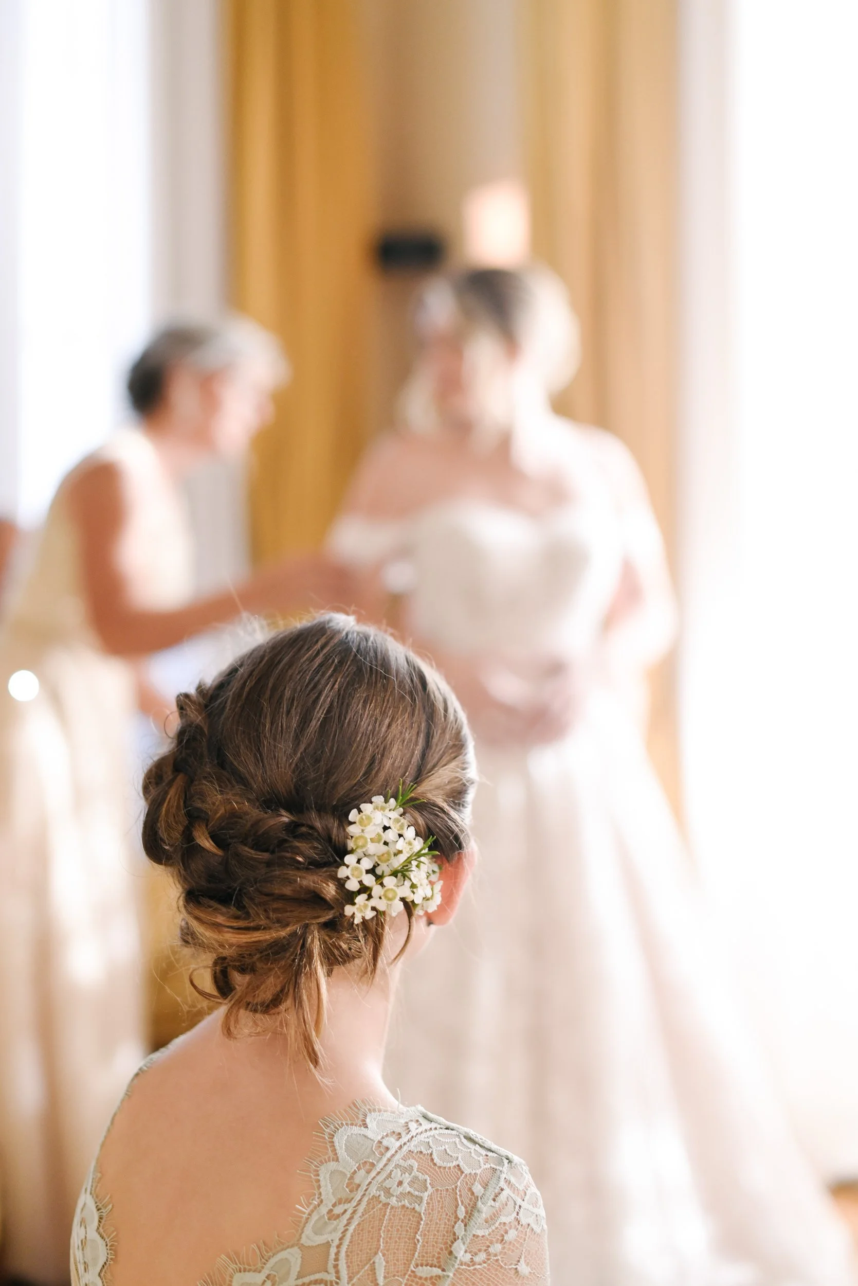 A woman with braided hair and white flowers in her hair, seen from behind, looking at another woman in a white dress in a well-lit room with curtains.