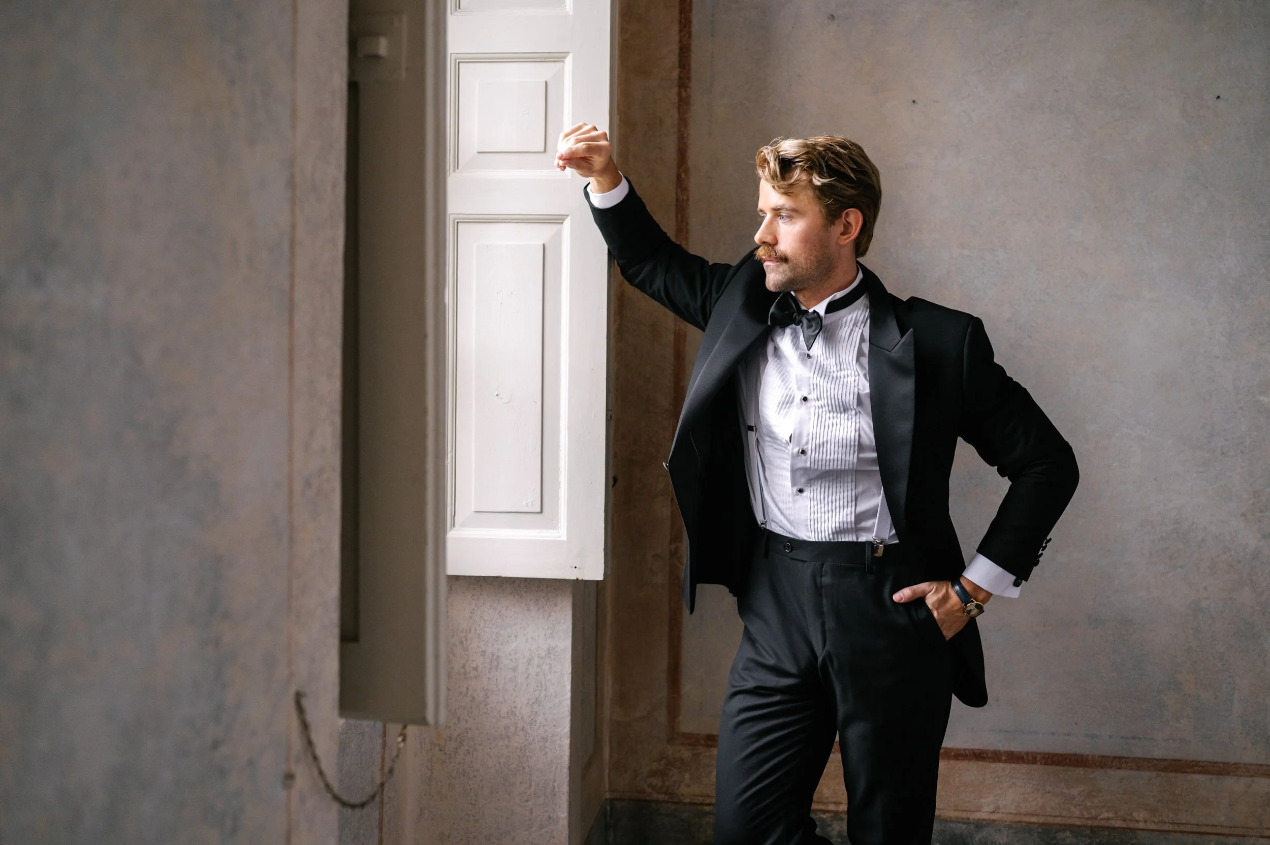 A man in a tuxedo with a bow tie posing confidently in front of a textured wall, with one hand in his pocket and the other resting on a white cabinet.