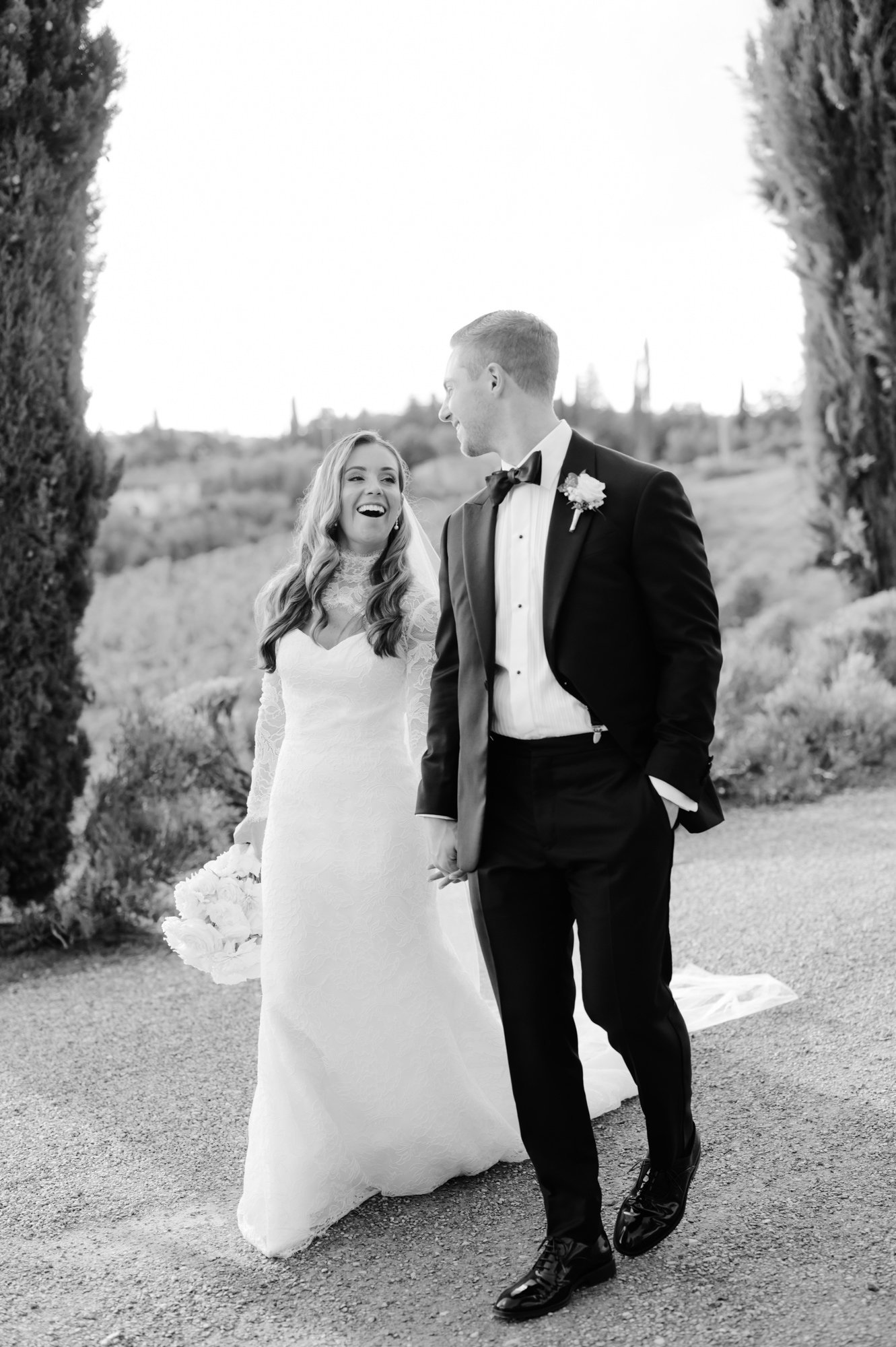 A bride and groom walking outdoors in wedding attire, smiling and holding hands, with trees and landscape in the background.