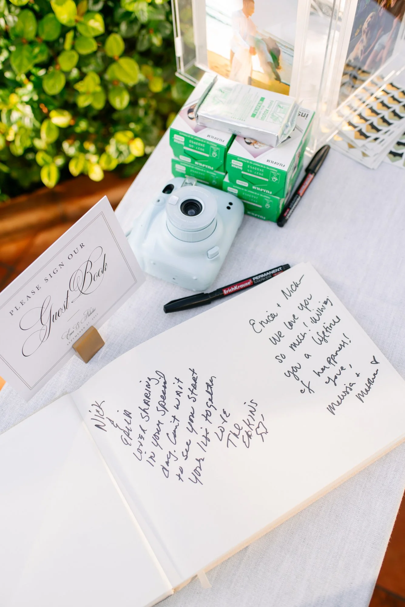 A guest book table at a wedding with a sign asking guests to sign the guest book, a handwritten note from the bride and groom, a white instant camera, stacks of film, and a few pens.
