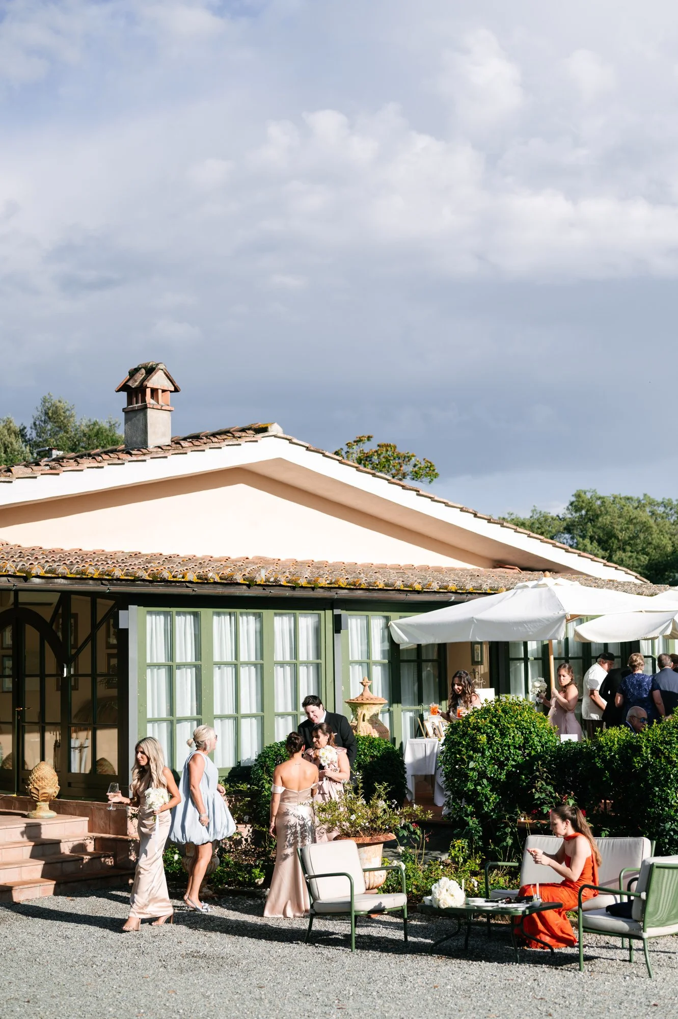 People attending an outdoor party or wedding reception near a house with a garden and patio furniture, under a cloudy sky.
