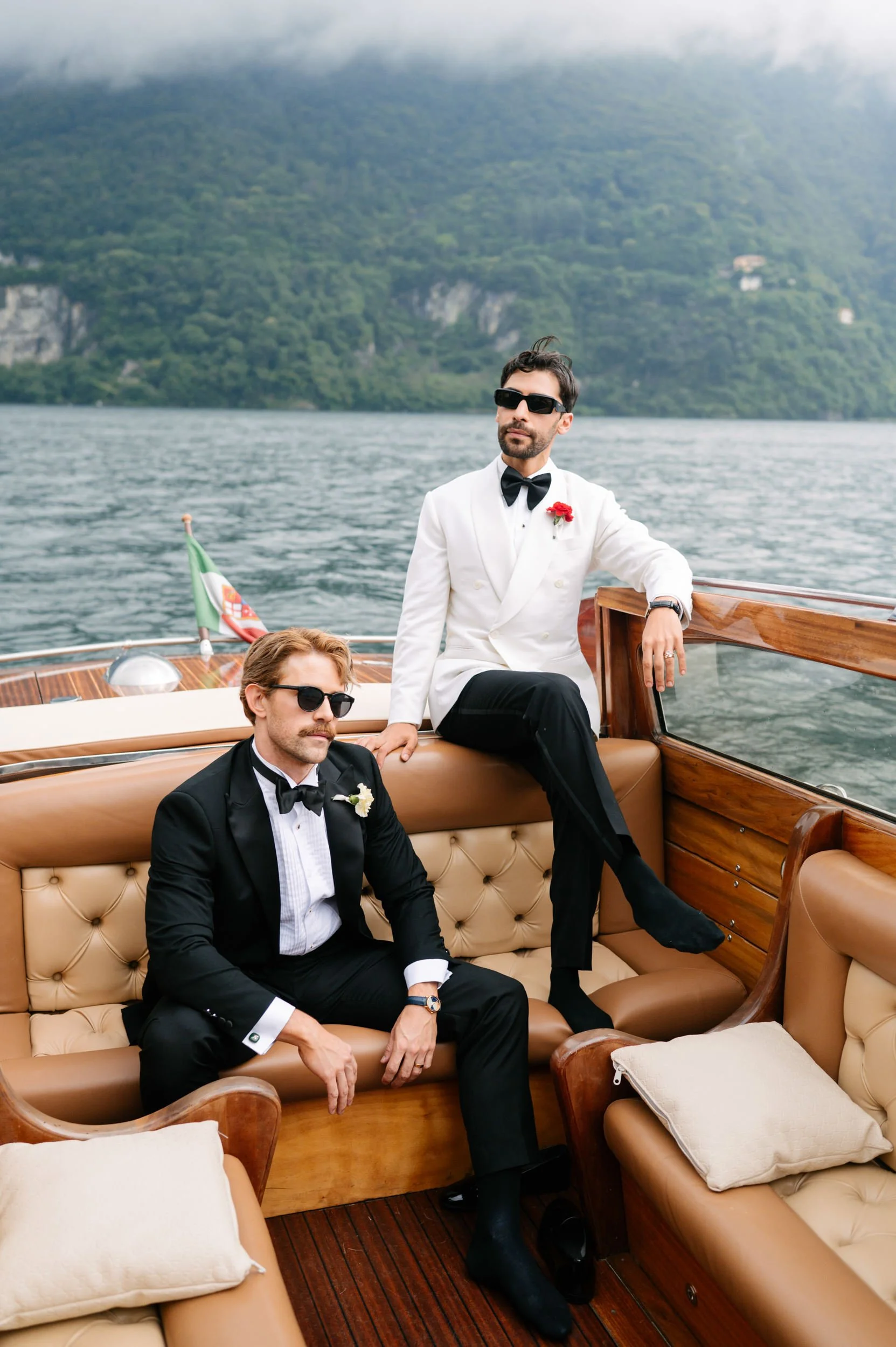 Two men dressed in tuxedos with sunglasses on a boat with a scenic lake and mountains in the background.