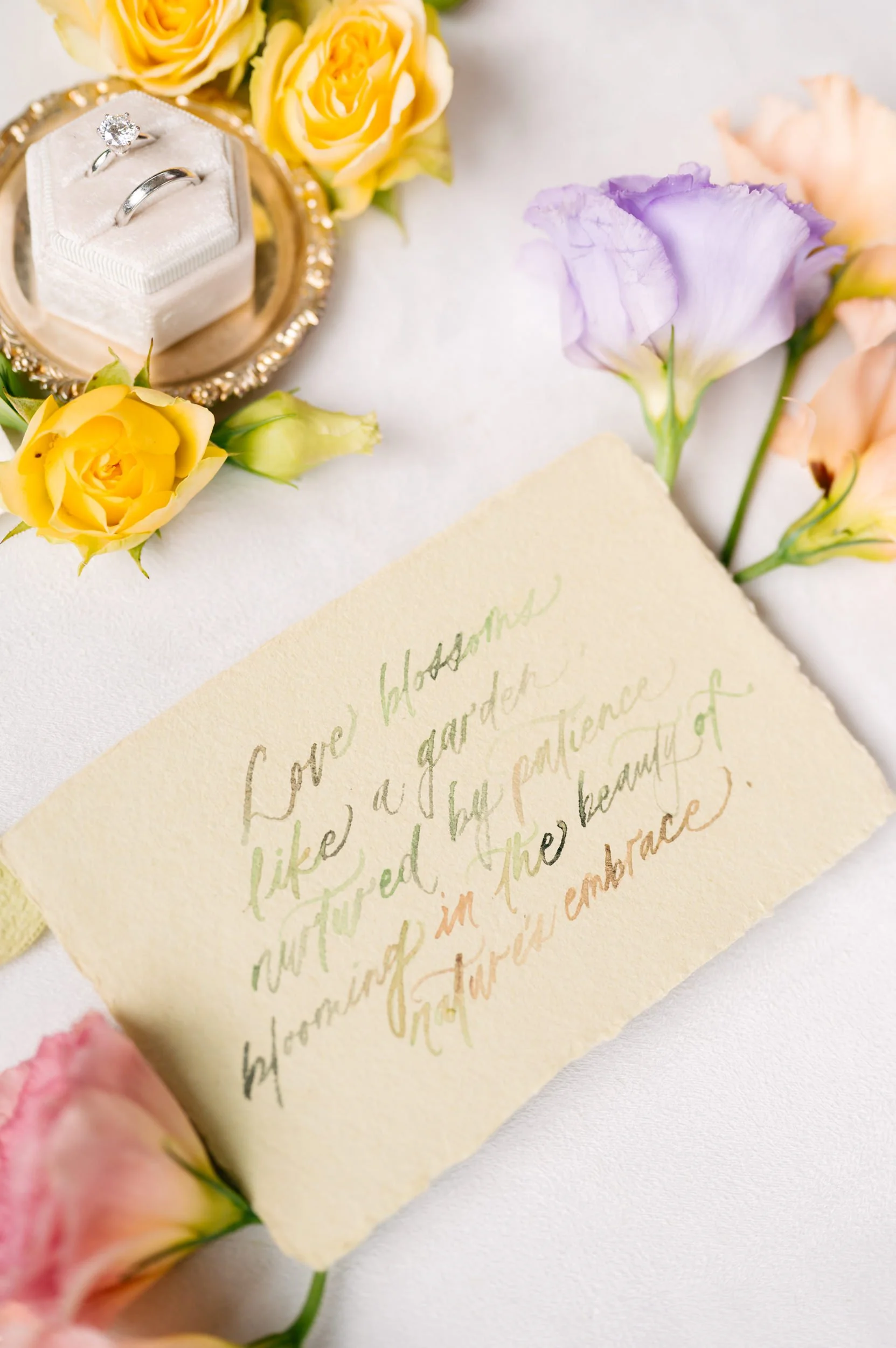 A small, white box with a diamond engagement ring and matching wedding band on a velvet cushion, surrounded by yellow and pale pink roses and a handwritten note on textured paper.