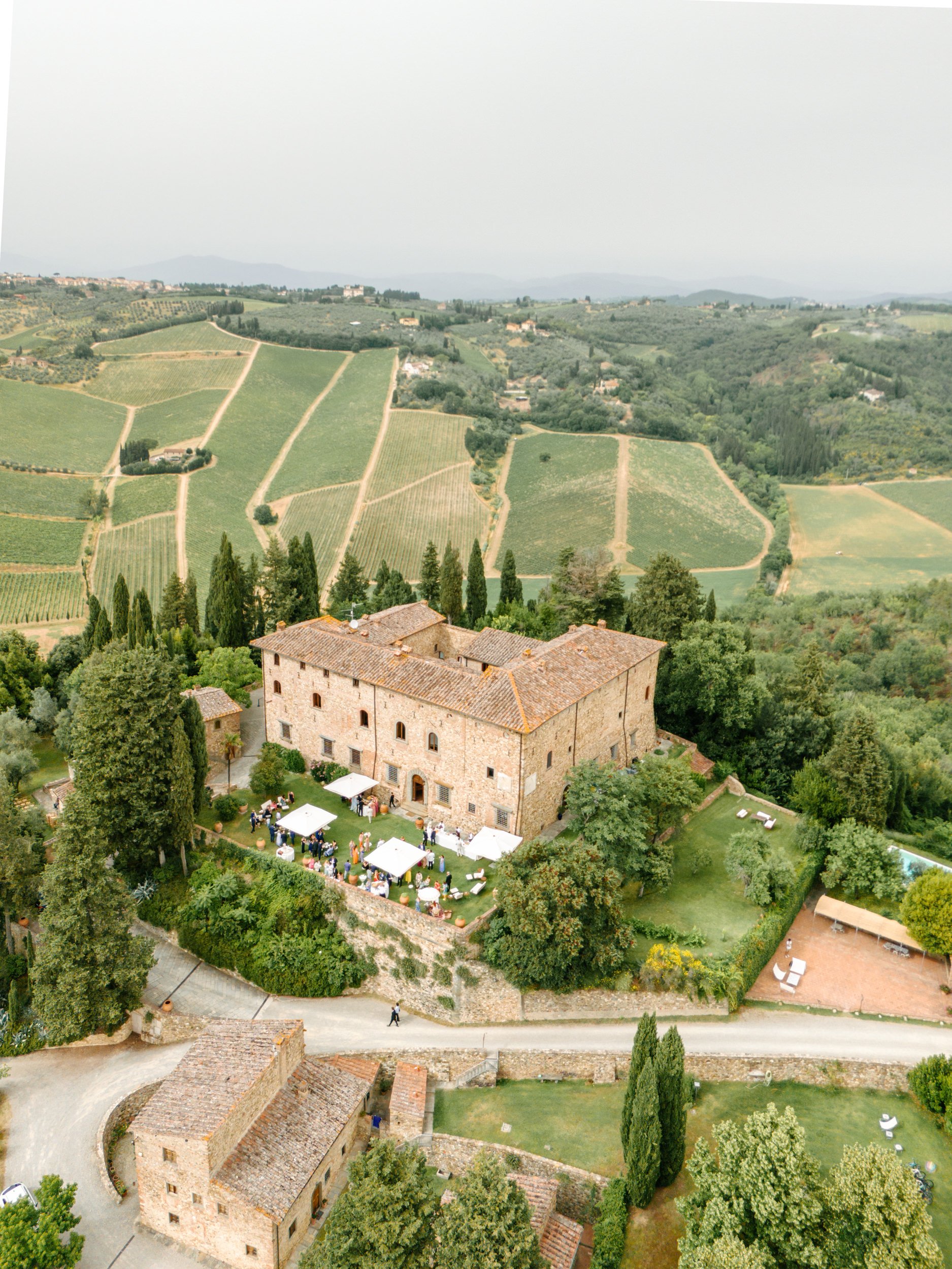 Aerial view of a historic stone building with outdoor event setup, surrounded by lush greenery, trees, and rolling green hills with vineyards in the background.