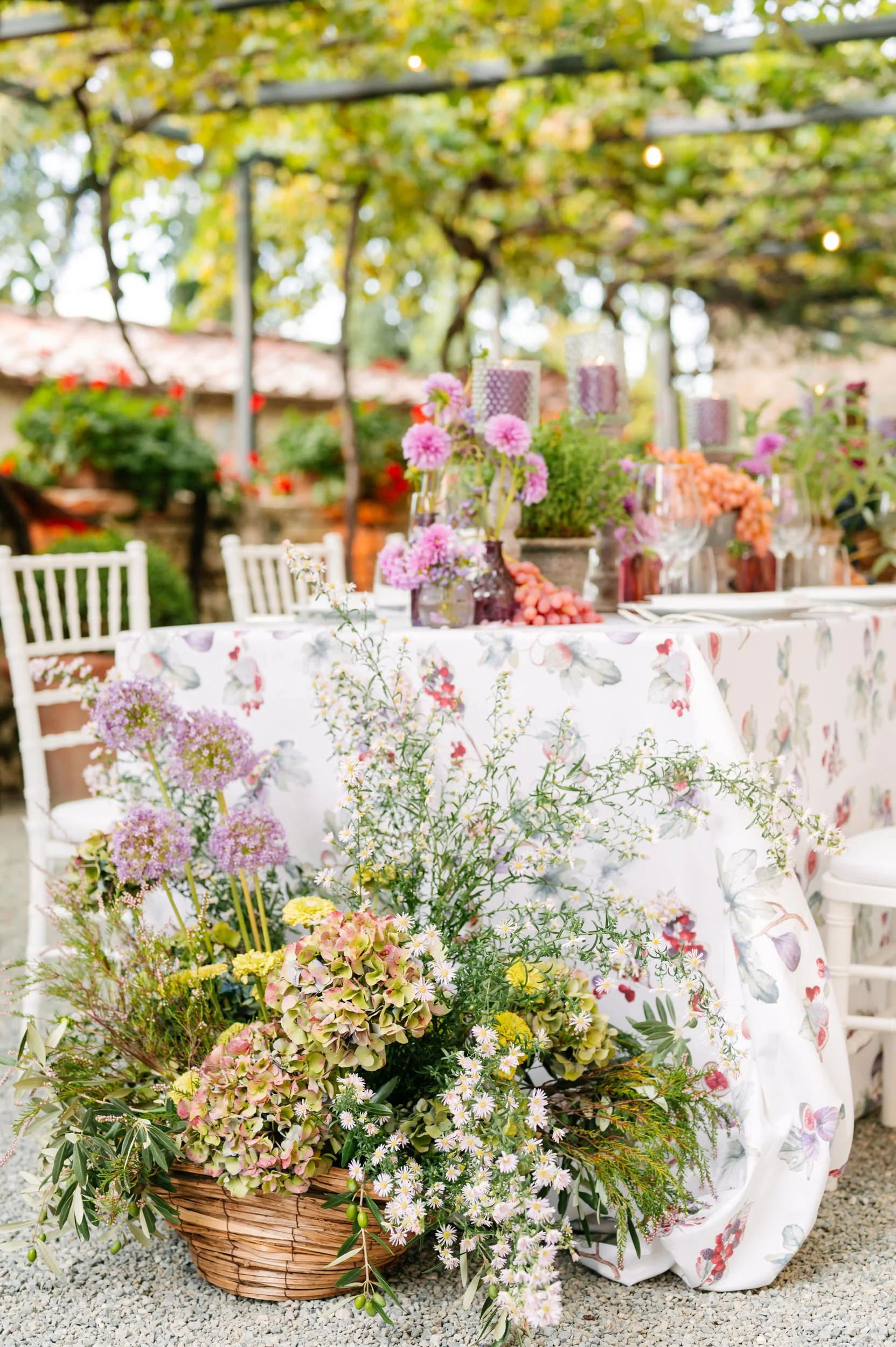 A beautifully decorated outdoor table with a floral tablecloth, surrounded by white chairs, adorned with pink, purple, and orange flowers in glass vases, candles, and additional flower arrangements, set in a garden with trees and greenery.