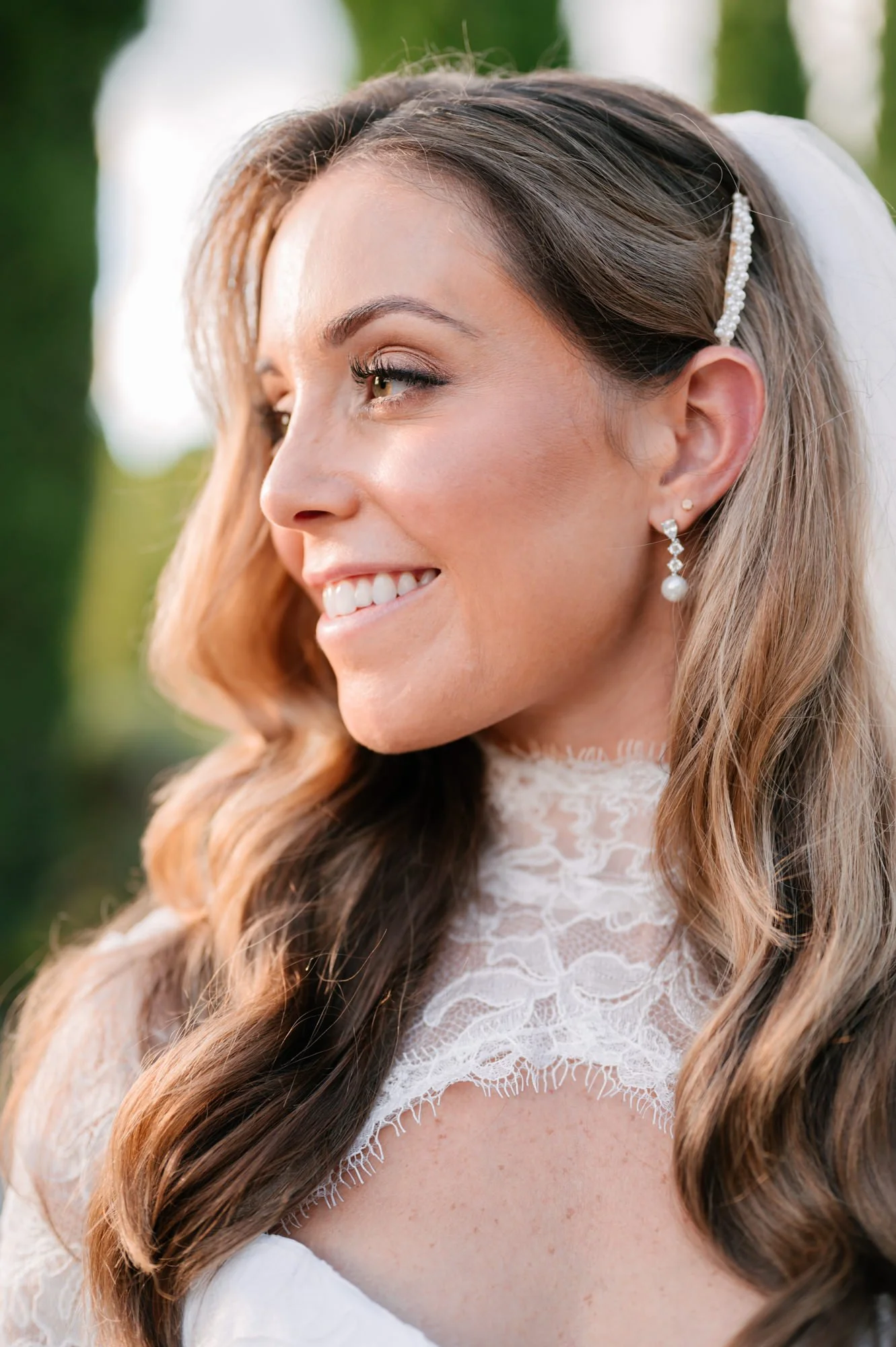 Close-up of a smiling woman in a white lace dress, with long wavy hair and pearl earrings, outdoors during daylight.