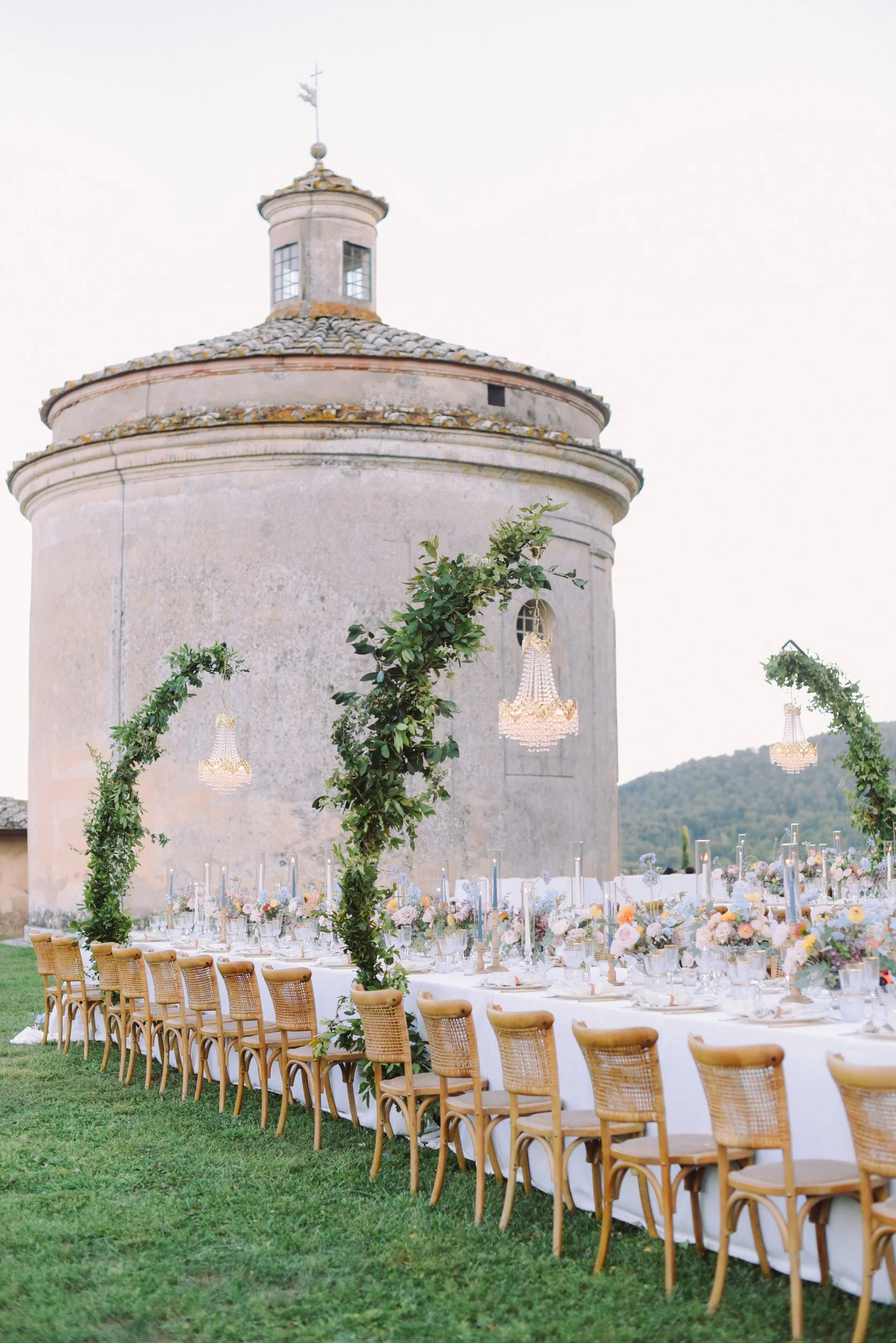 An outdoor banquet table set up in front of an old stone building with a round tower and a weather vane on top, decorated with flowers, greenery, and hanging chandeliers.
