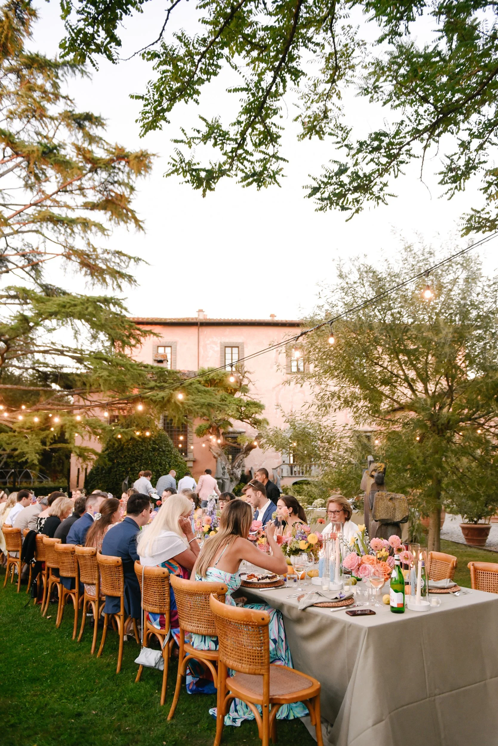 Outdoor dinner party with guests seated at a long table decorated with pink flowers, surrounded by trees and string lights, at sunset.