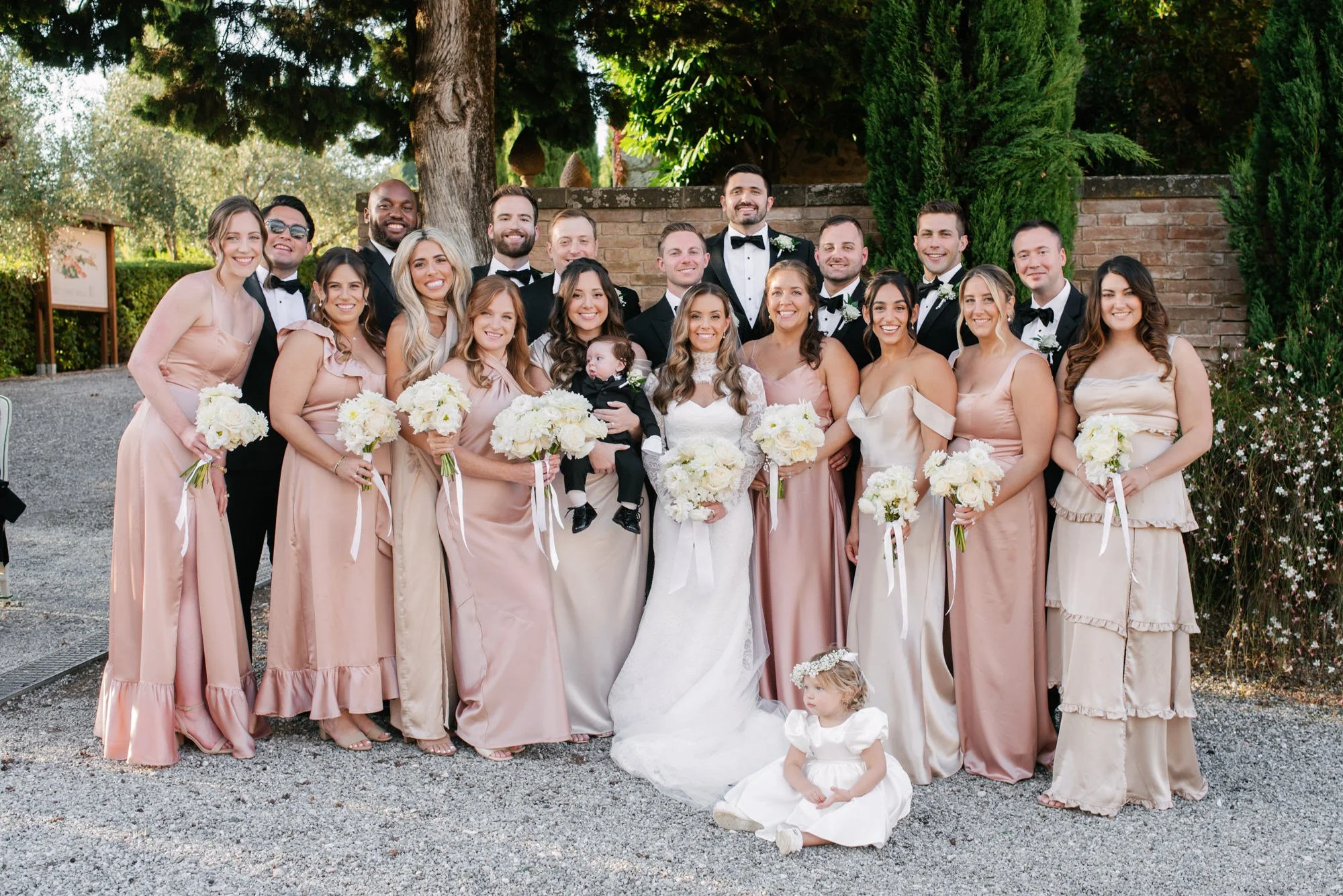 Group of people at a wedding, including the bride in a white dress, bridesmaids in blush dresses, groomsmen in tuxedos, and children, outdoors under trees with greenery in the background. Luxury editorial wedding photography in Tuscany. 