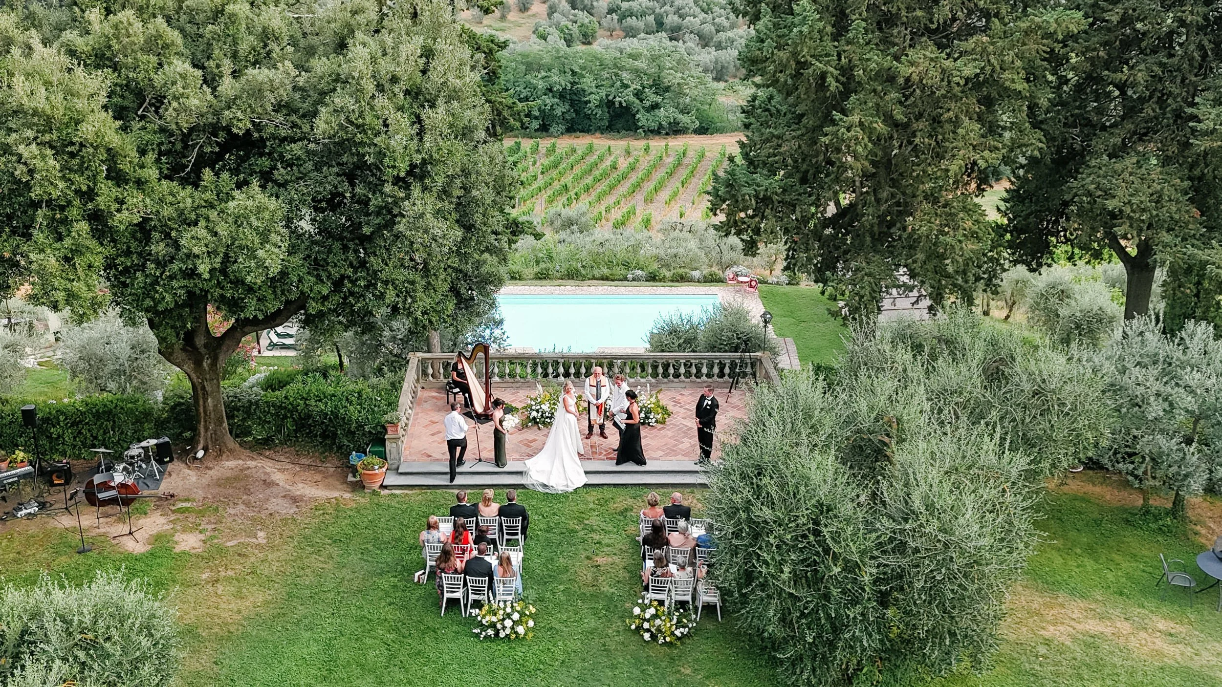 An outdoor wedding ceremony in a scenic garden with a small stage, trees, a swimming pool, and guests seated on white chairs. The bride and groom stand near the officiant.