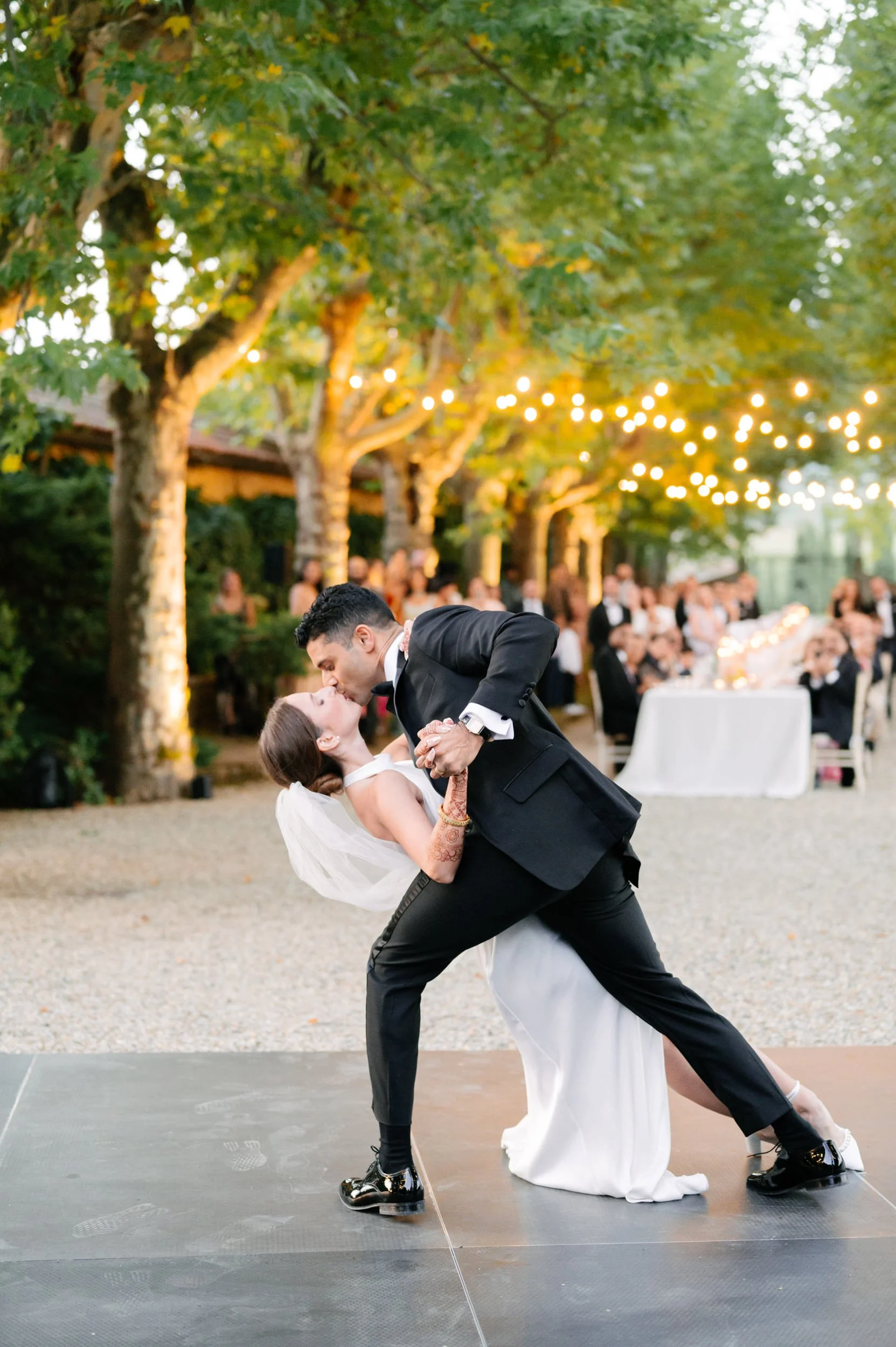 A bride and groom share a dance at an outdoor wedding reception under string lights, with guests seated at tables in the background.