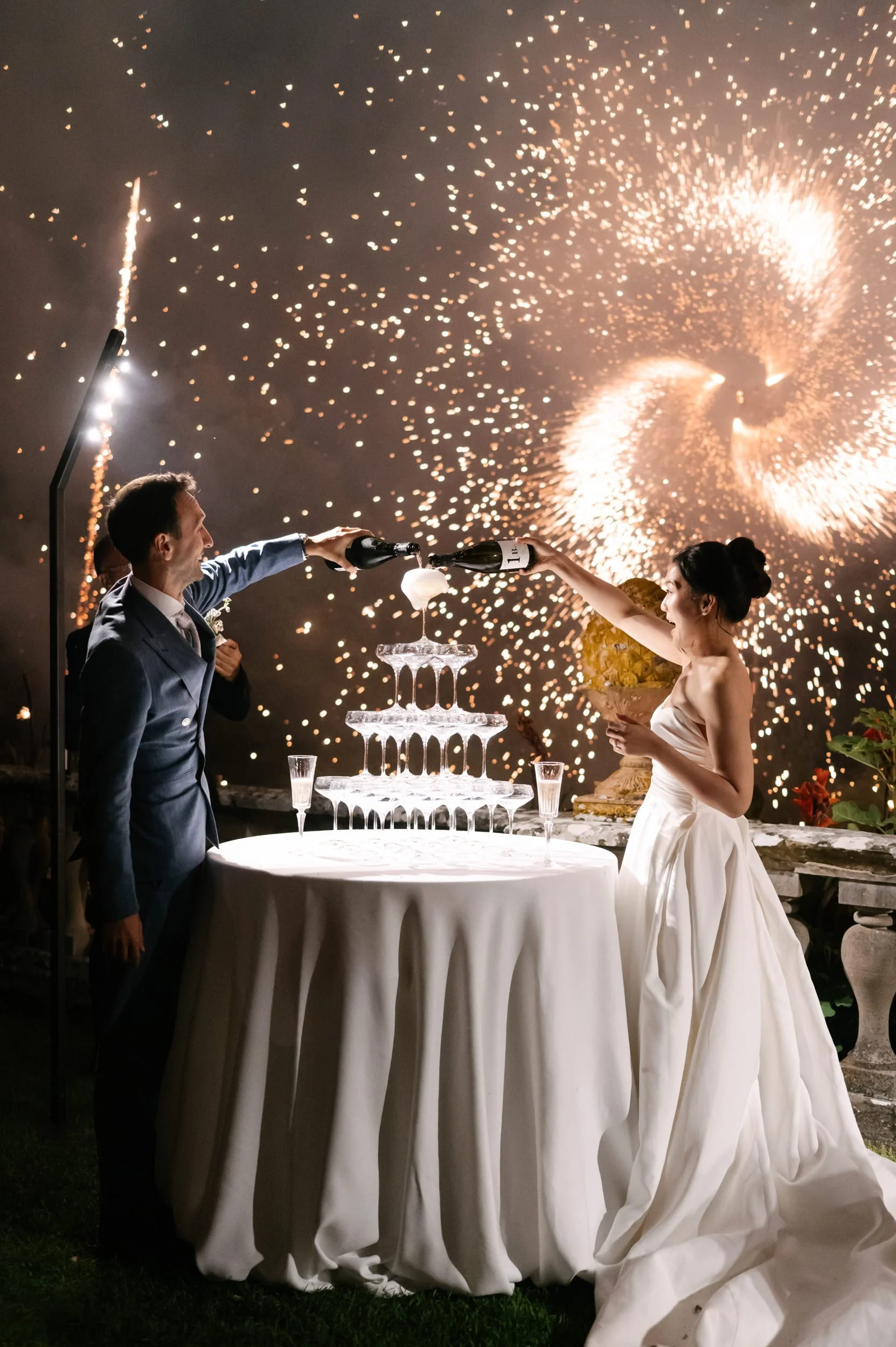 A couple, dressed in wedding attire, celebrating by pouring champagne into a tower of glasses at their wedding reception, with fireworks in the background.