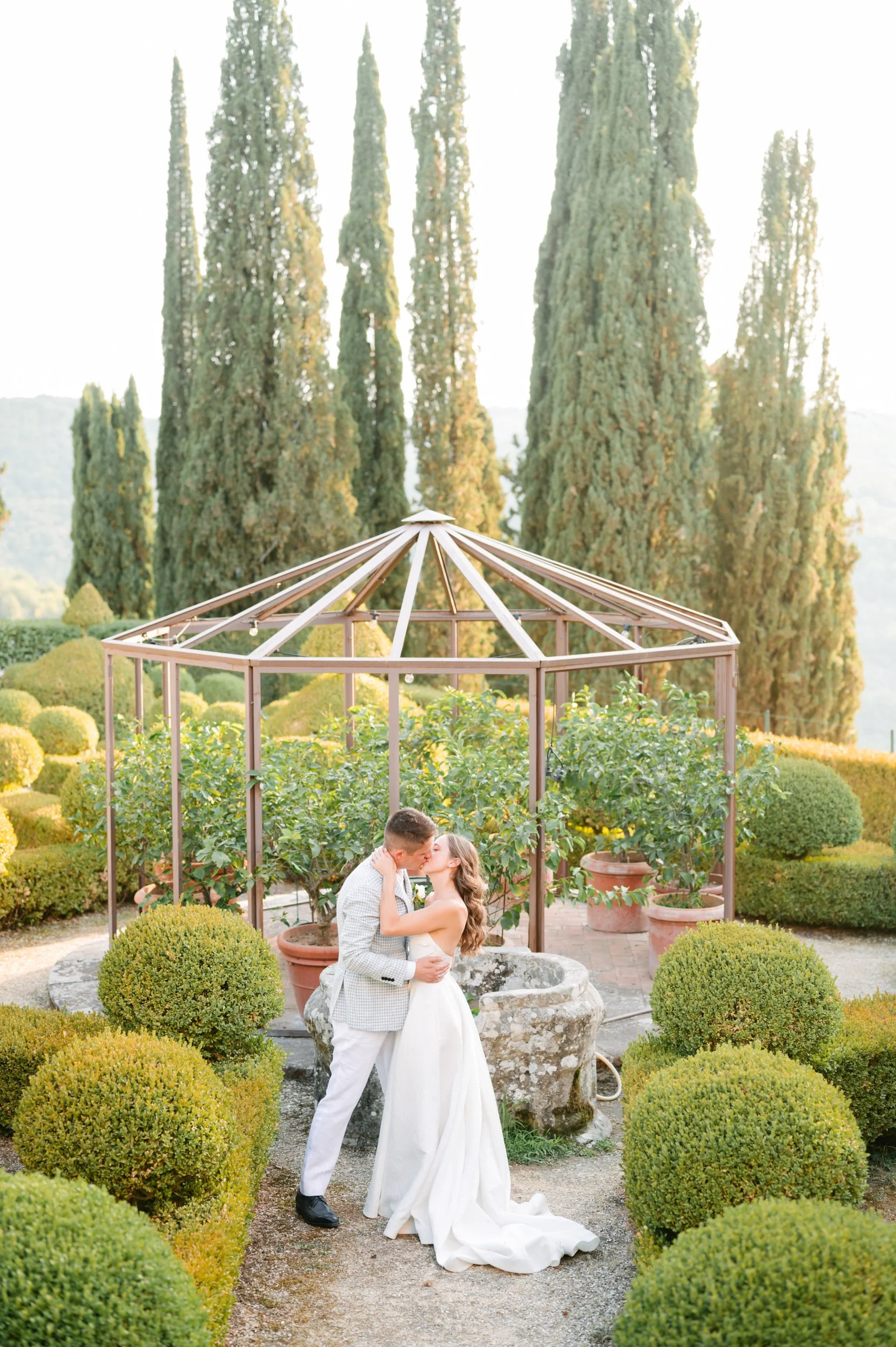 A bride and groom kissing in a lush garden with tall cypress trees and trimmed bushes, standing near a stone fountain under a gazebo.