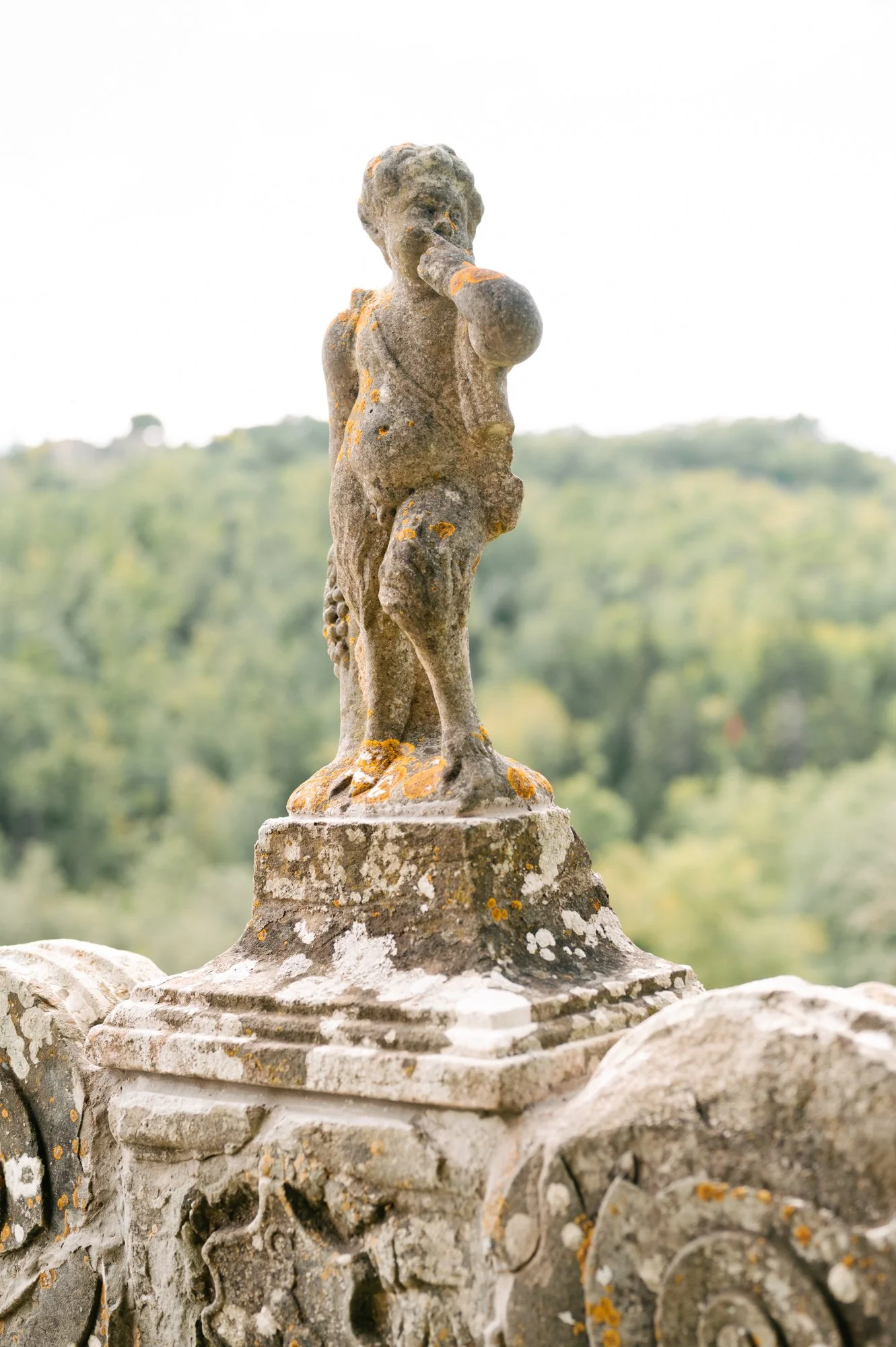 A weathered stone cherub statue on a decorative stone fence with a blurred green landscape in the background.