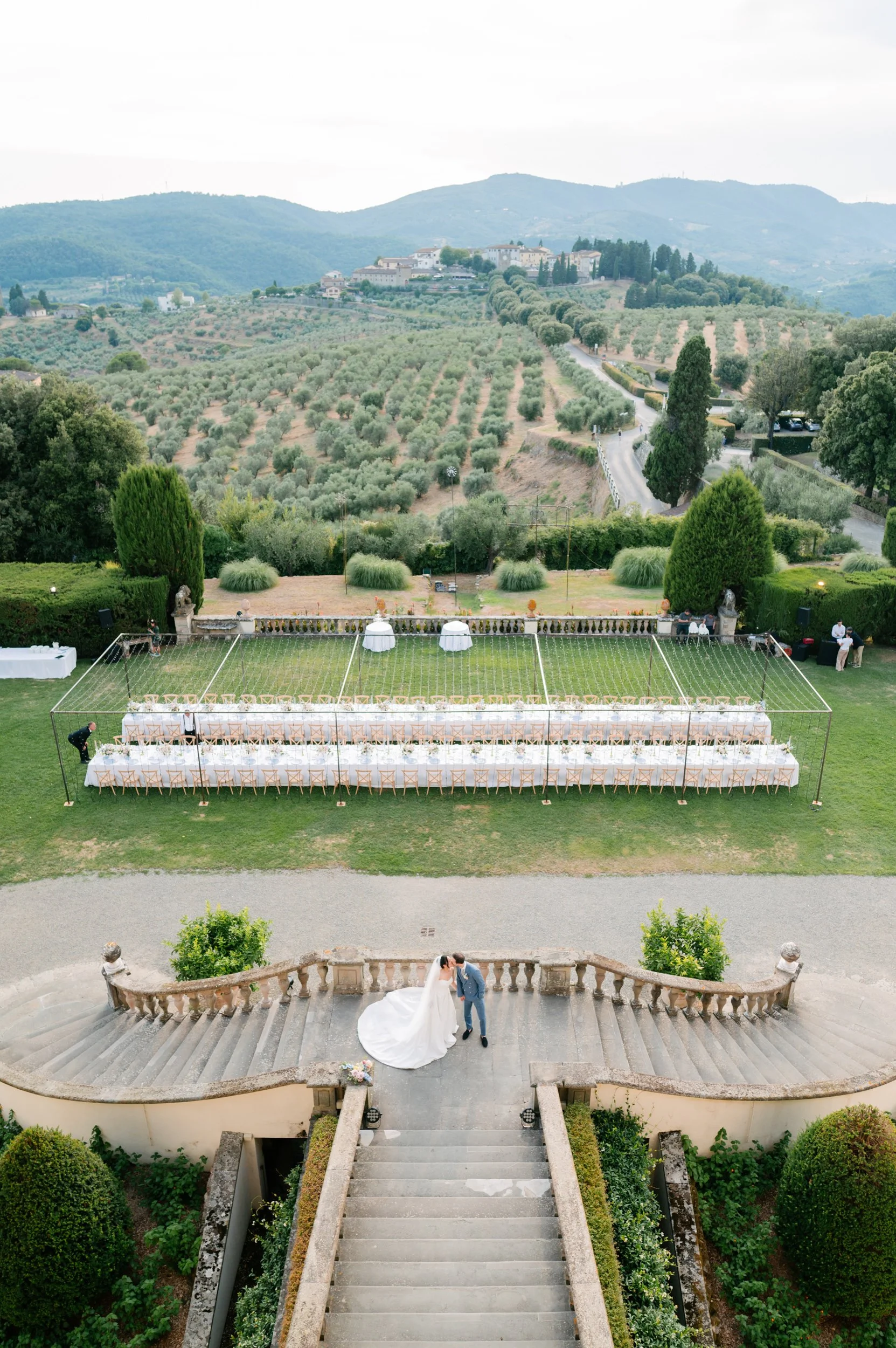 A wedding couple standing on the steps of an outdoor terrace overlooking a large, decorated event space with long tables, set for a wedding reception, and scenic rolling hills and vineyards in the background.