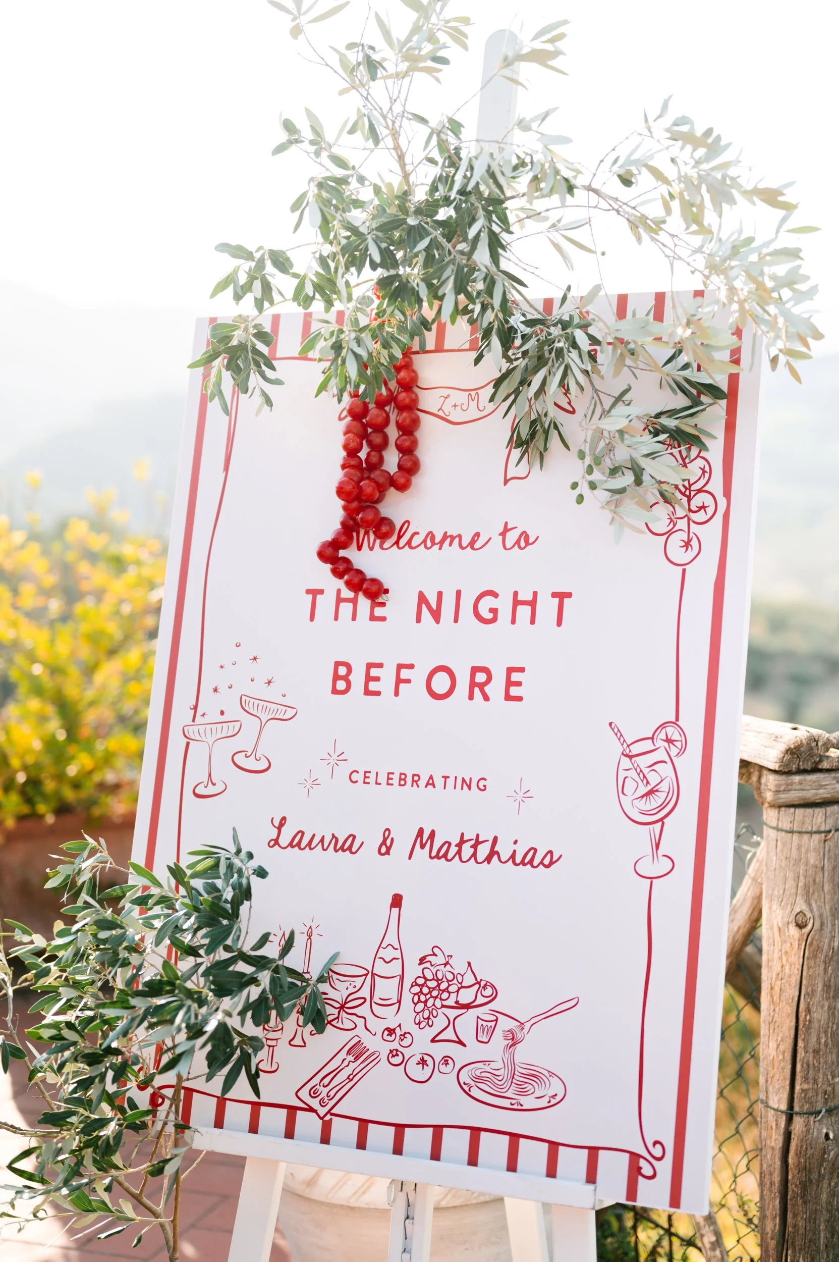 A decorative welcome sign for an event called 'The Night Before,' celebrating Laura and Matthias. The sign is decorated with greenery and a string of red berries, with illustrations of drinks, food, and celebration items at the bottom.