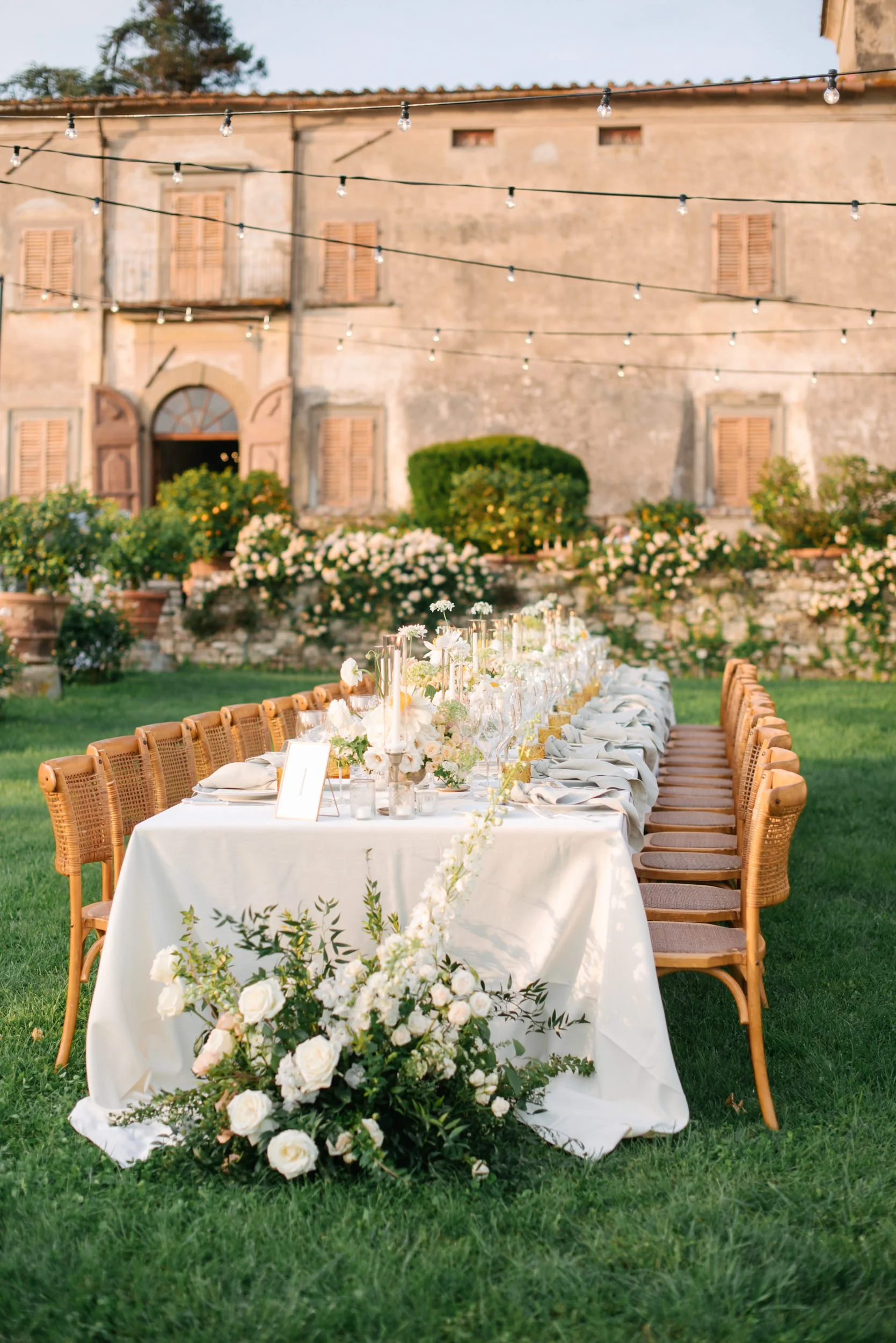 A long outdoor wedding or event table decorated with white flowers, candles, and glassware, set on a grassy lawn with an old rustic building and string lights overhead.