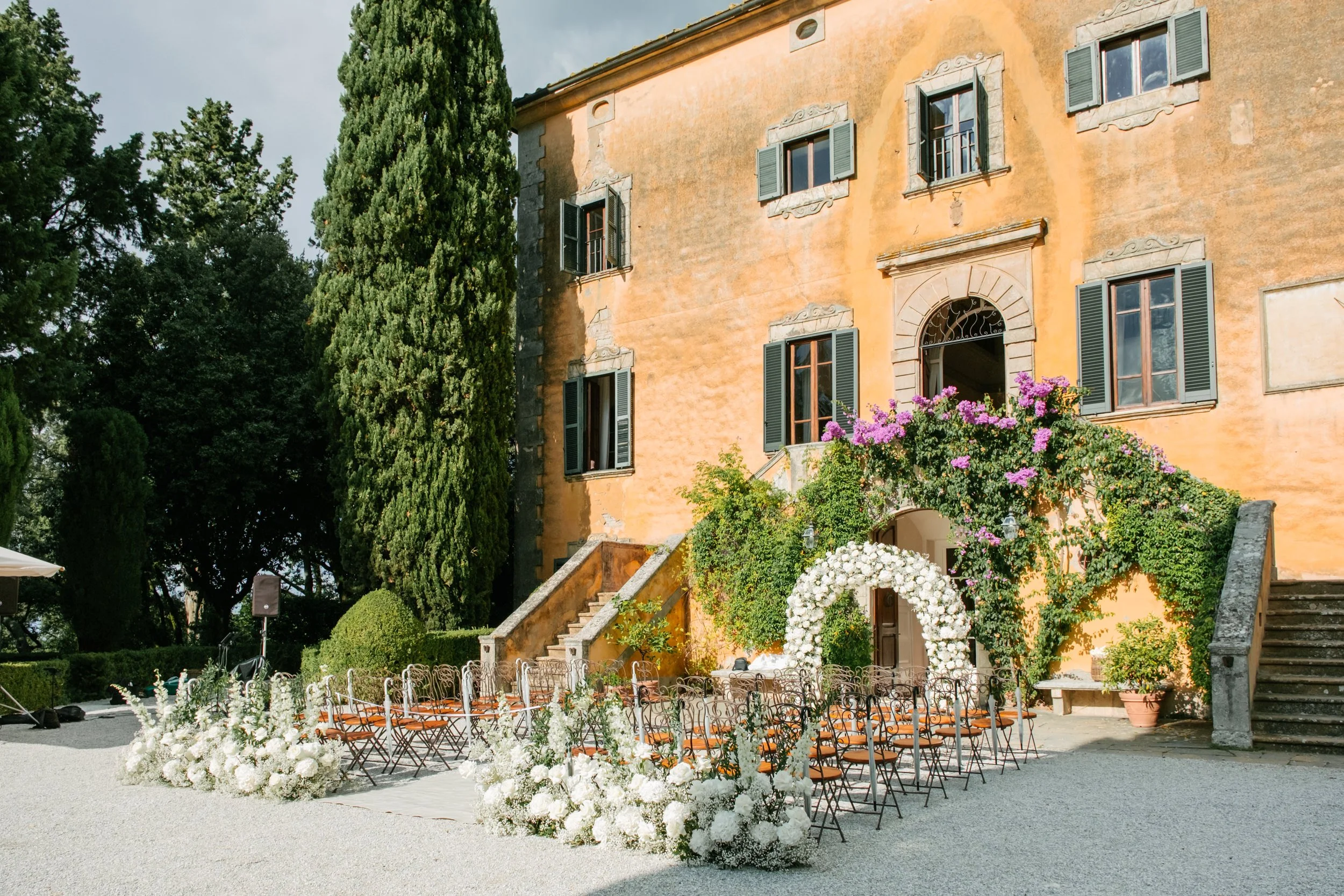 Outdoor wedding setup in front of an old yellow building with green shutters, surrounded by trees, with white floral arrangements, a floral arch, and chairs arranged for guests.