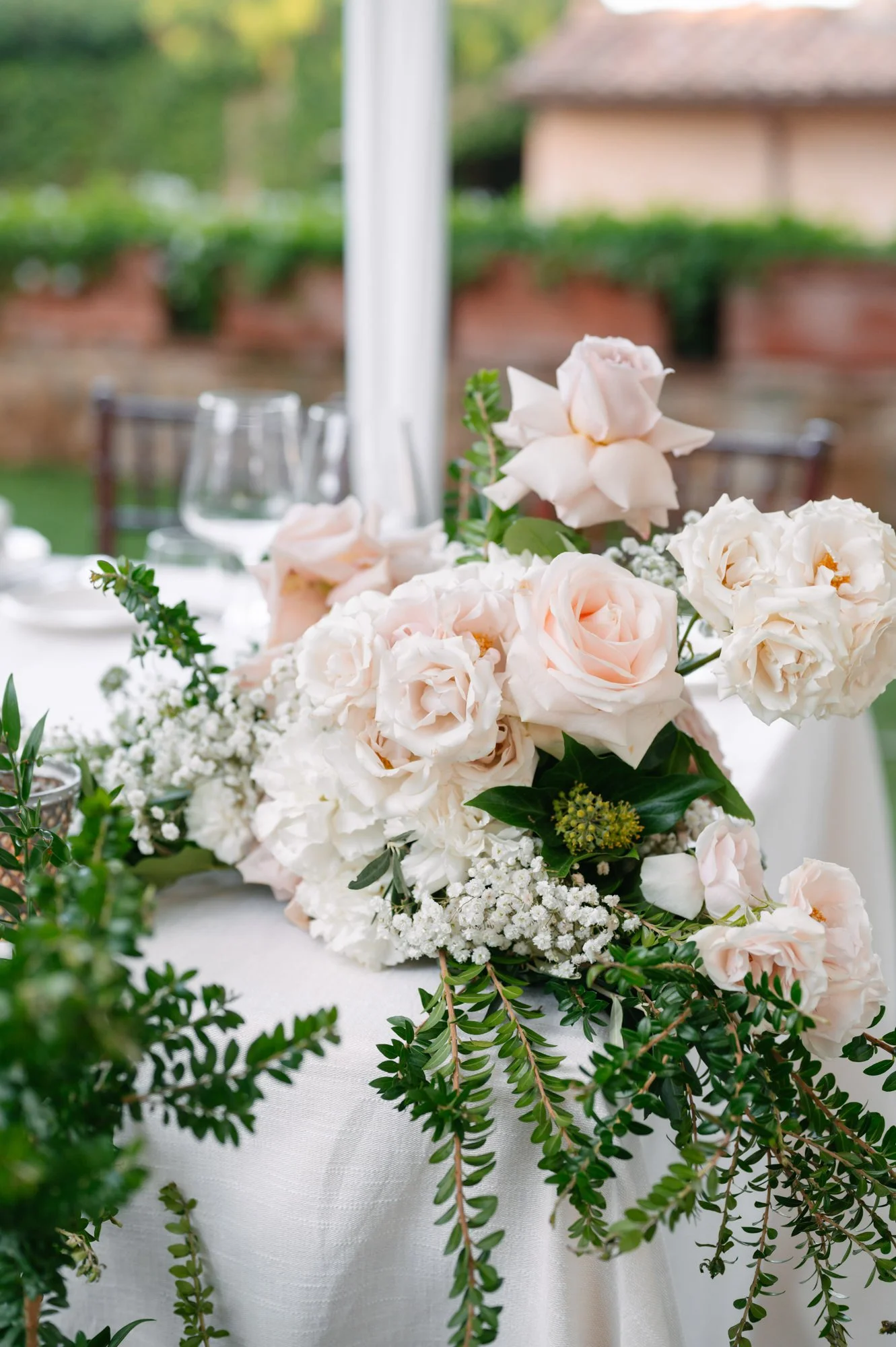 Elegant wedding table centerpiece featuring blush pink roses, white hydrangeas, baby's breath, and greenery on a white tablecloth with wine glasses and a blurred outdoor background.