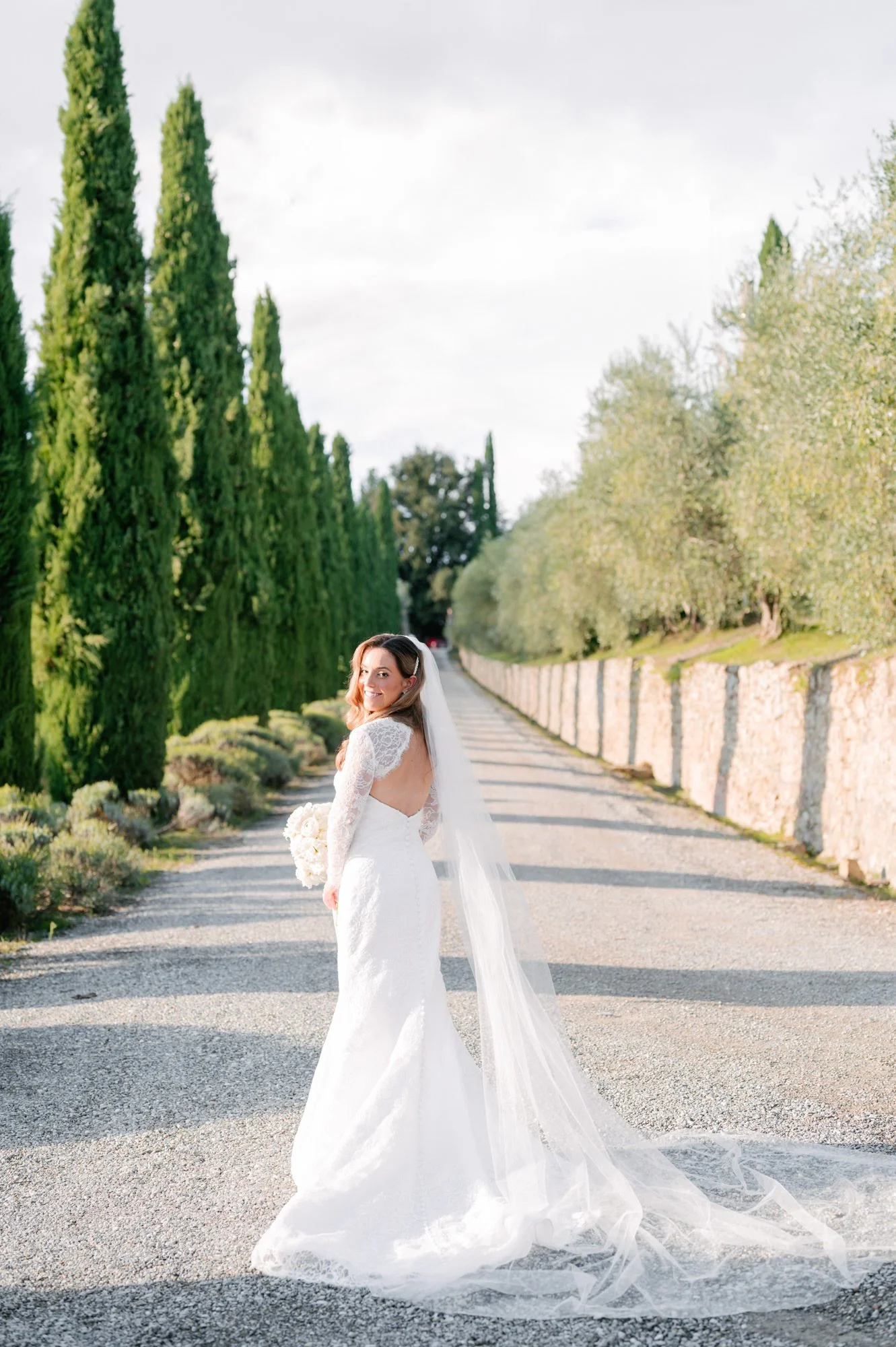 A bride in a white wedding dress holding a bouquet, standing on a gravel road lined with tall cypress trees and a stone wall, smiling at the camera.