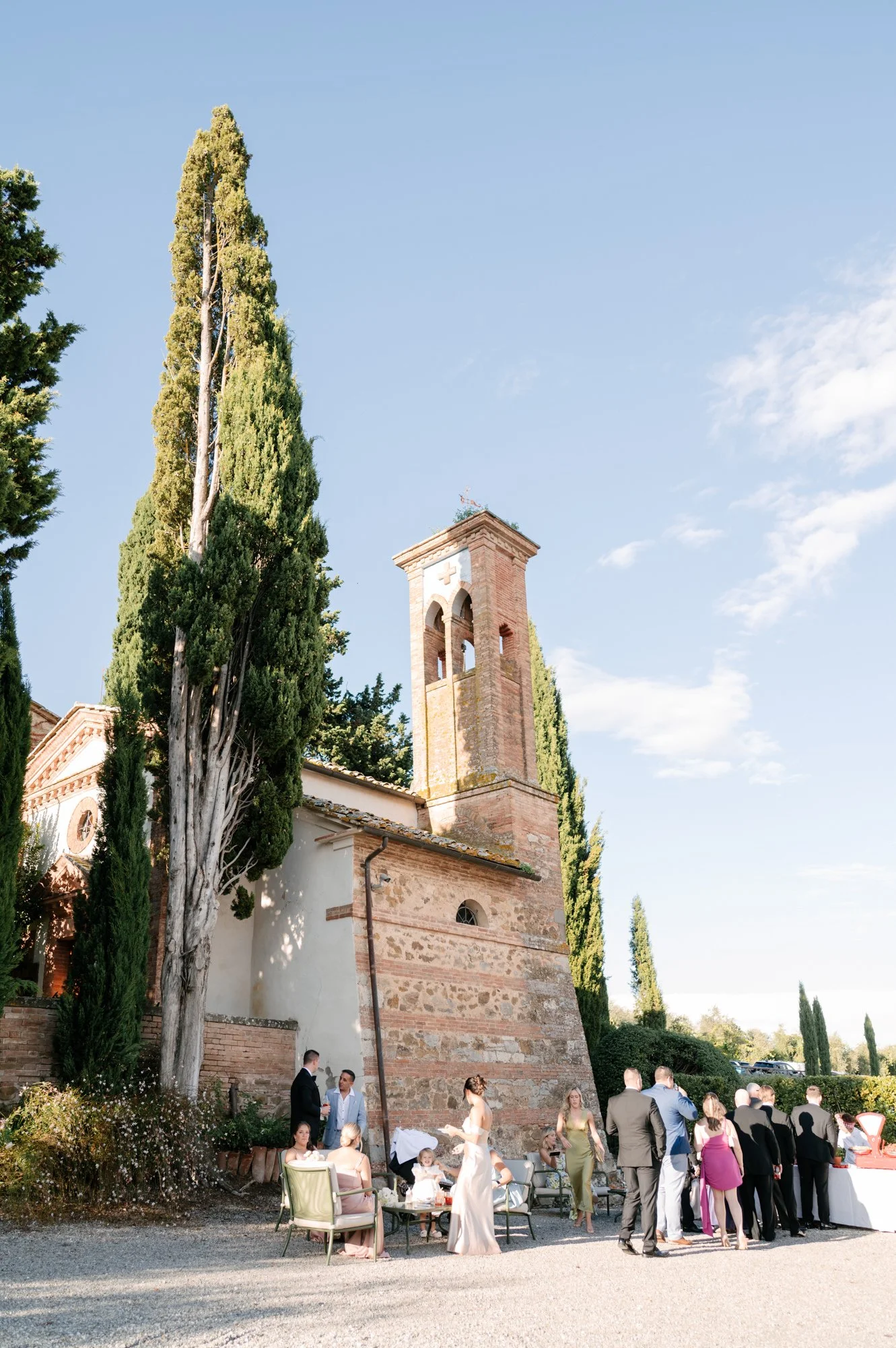A wedding reception gathering outside a historic brick church with tall cypress trees, where guests are socializing and enjoying the outdoor event.