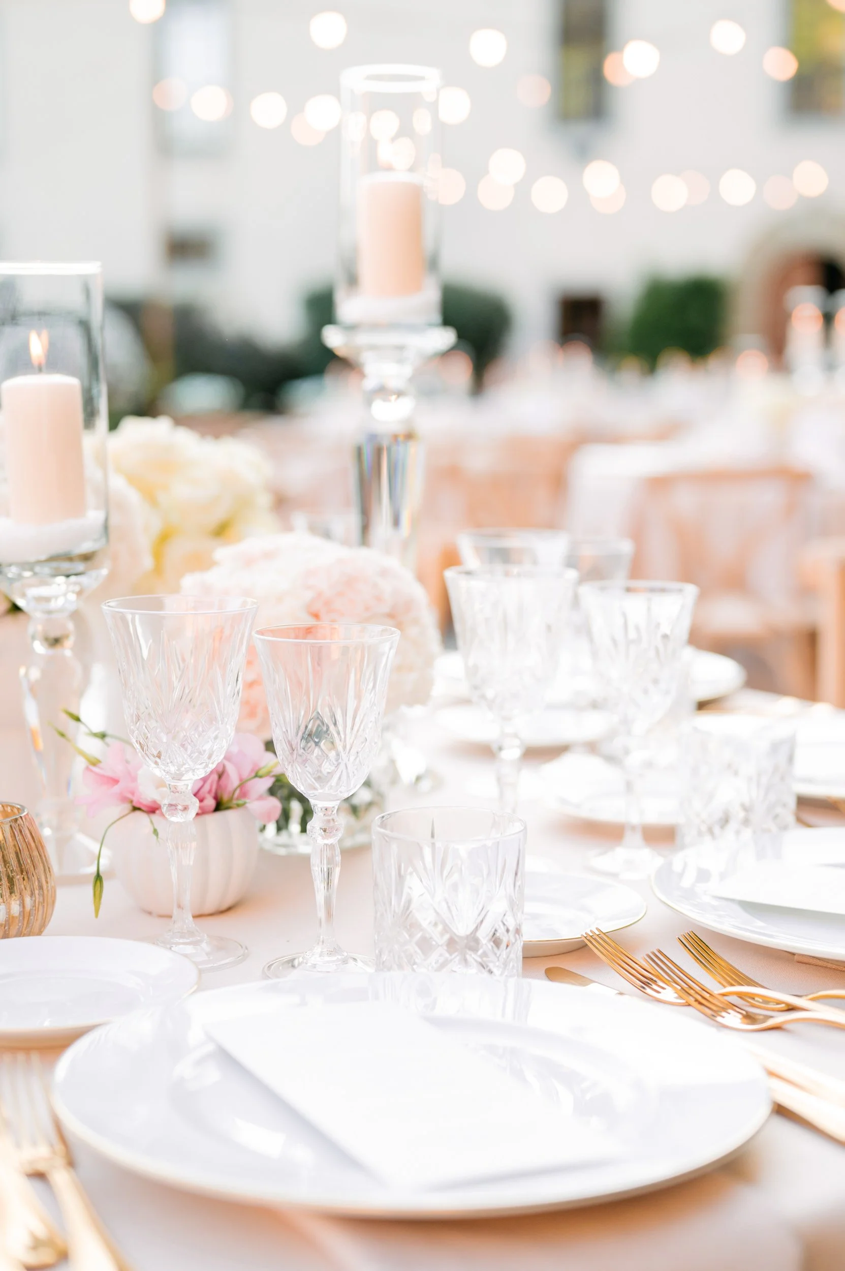 Elegant table setting with white plates, gold cutlery, and crystal glasses, decorated with pink and white flowers and tall candles, at an outdoor event.