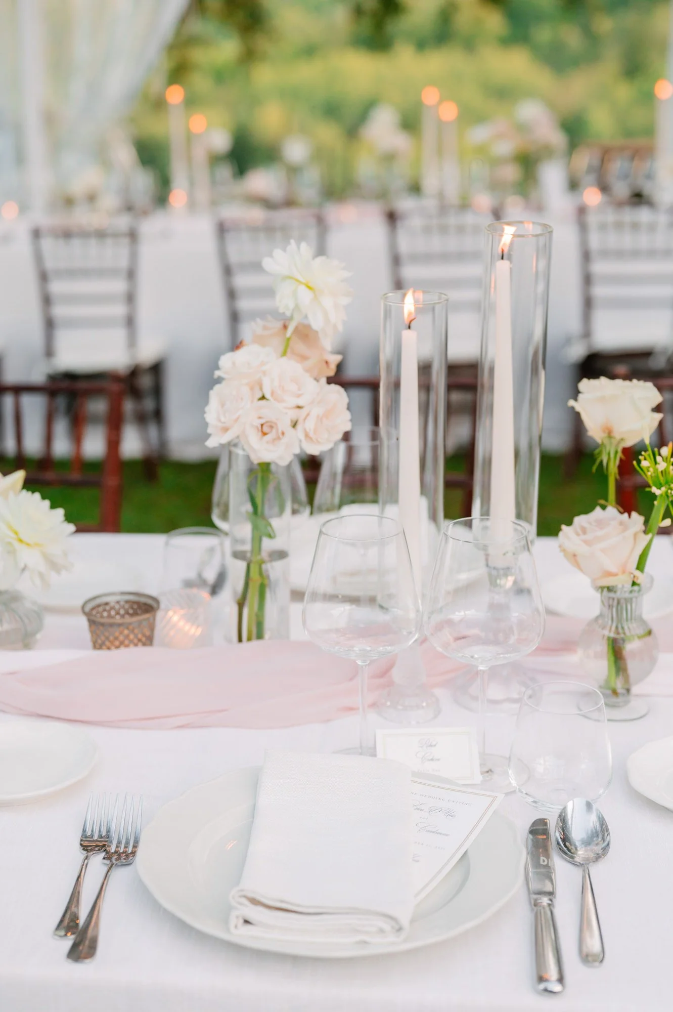 Elegant outdoor wedding table setting with pink flowers, candles, wine glasses, and white plates.