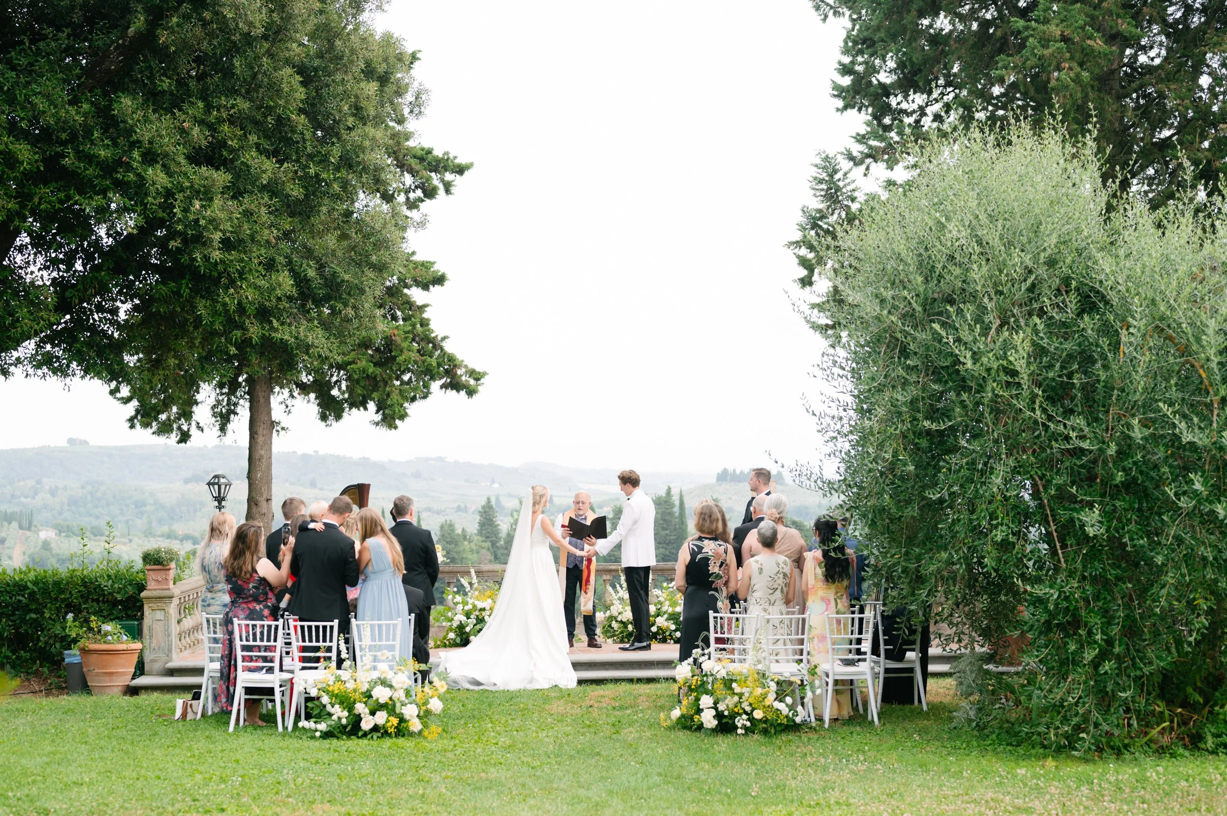 A wedding ceremony outdoors with a bride and groom standing on a platform, holding hands and reciting vows. The bride wears a white wedding gown, and the groom is in a white jacket. Wedding guests are seated and standing around, watching the couple. 