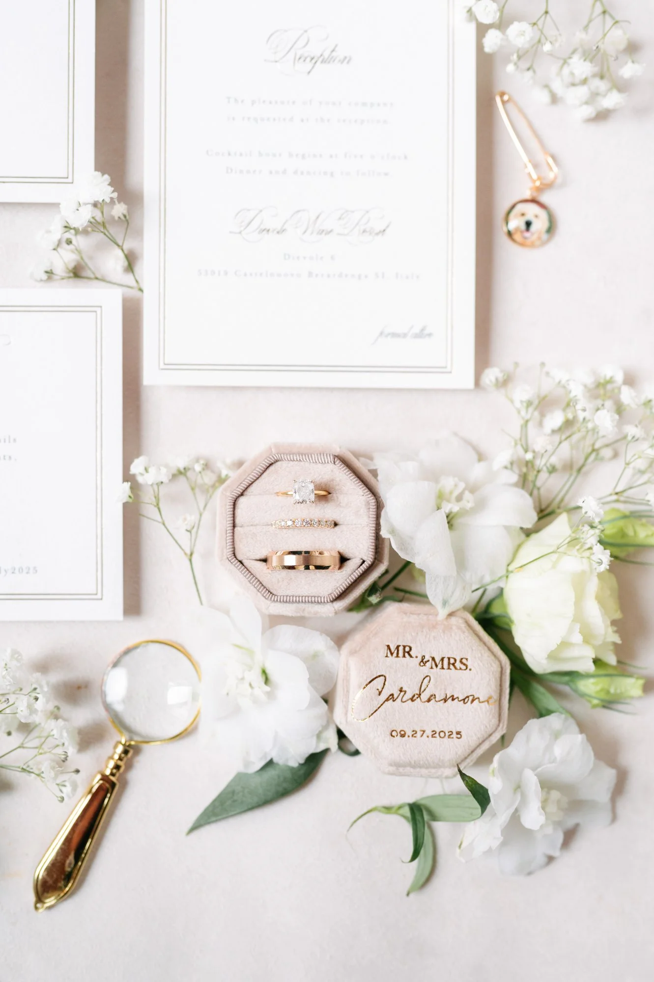 Wedding invitation, engagement rings in a jewelry box, boutonniere, magnifying glass, and white flowers arranged on a light surface.