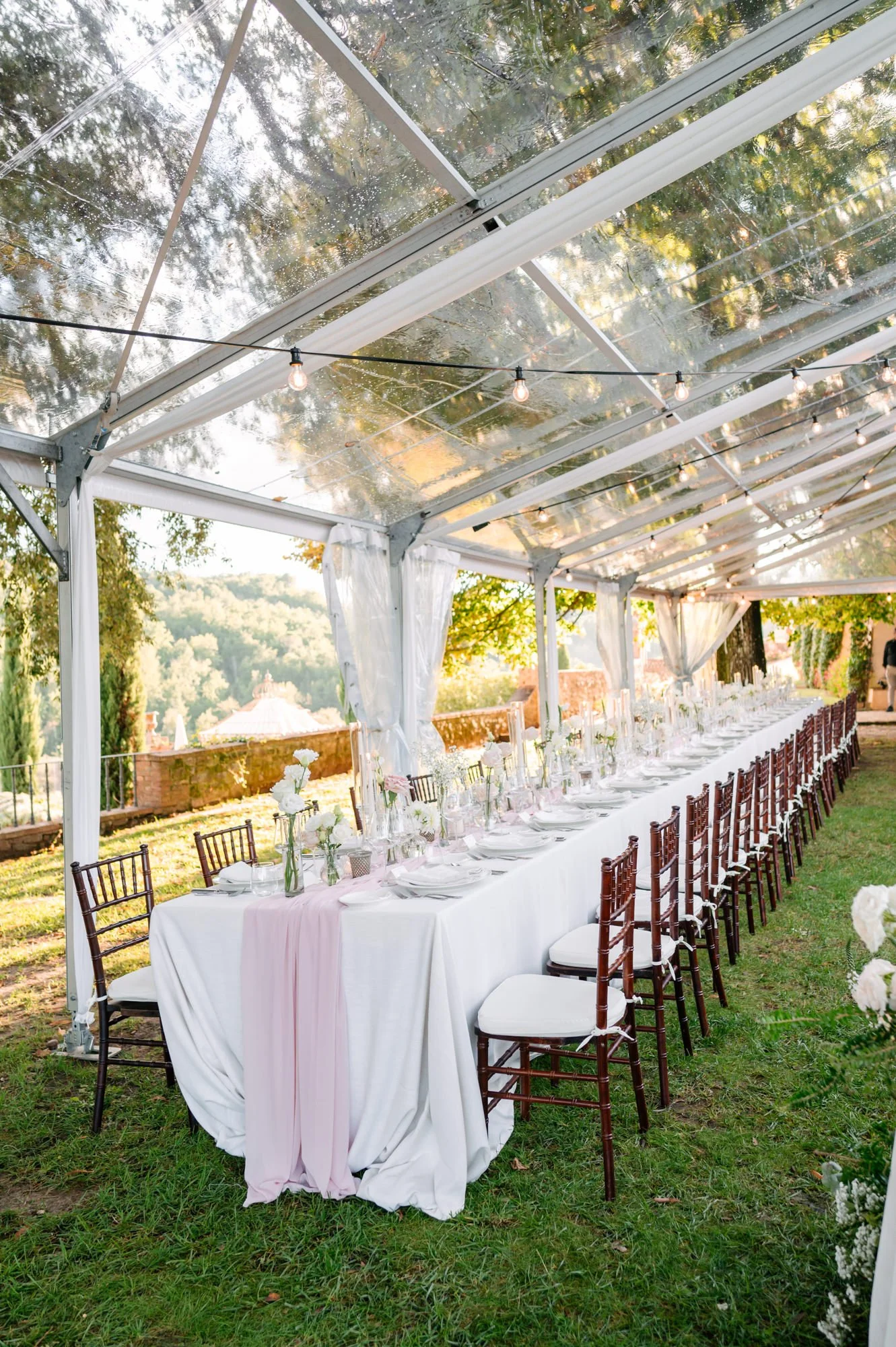 Long banquet table under an outdoor tent with string lights and white curtains, set up for an event with white tablecloths, floral centerpieces, and wooden chairs on a grass lawn with trees and a scenic view in the background.