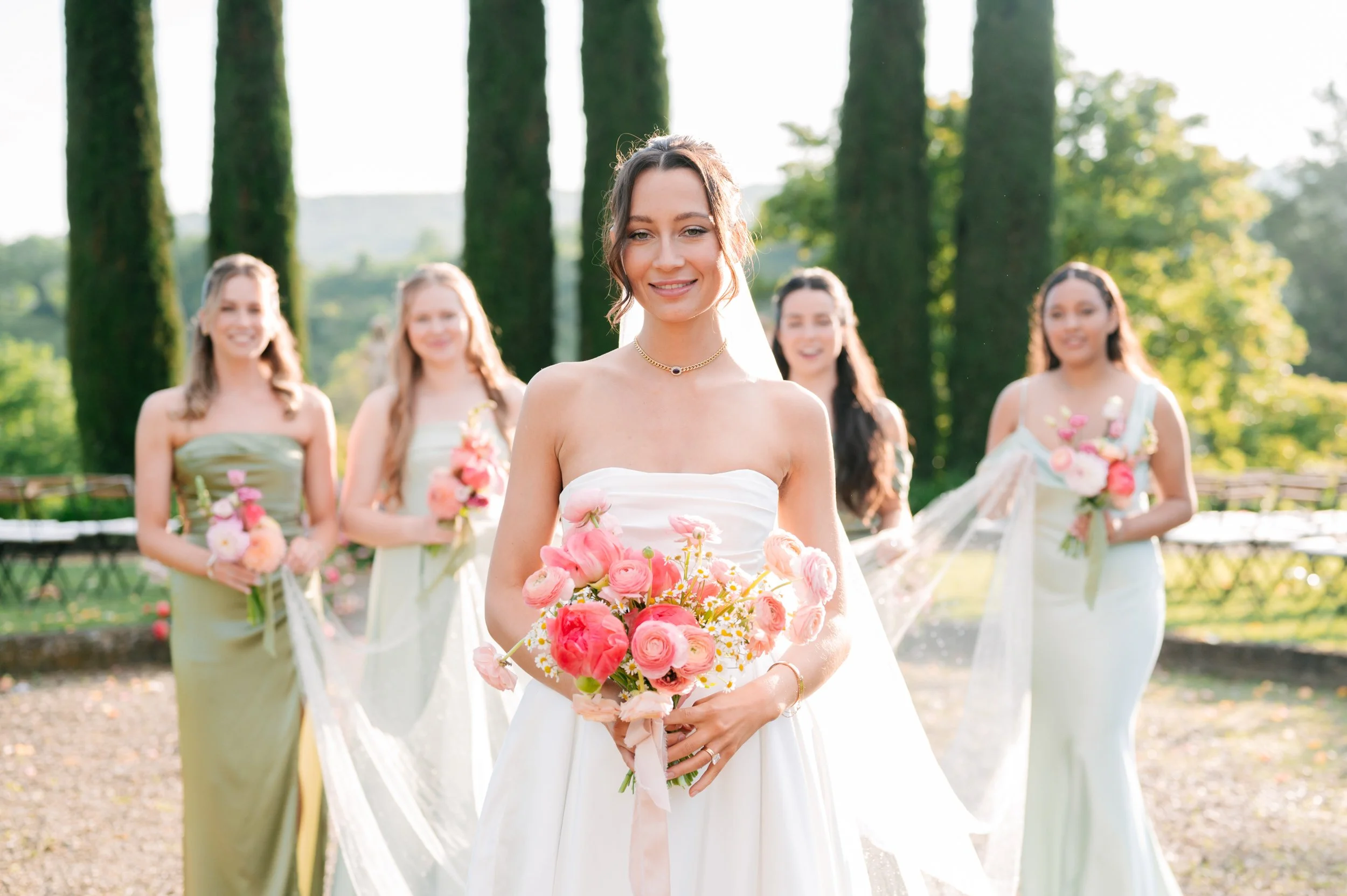 A bride in a white wedding dress holding a bouquet of pink flowers, standing outdoors with four bridesmaids in pastel dresses holding flowers, trees and green landscape in the background, bright and sunny setting.