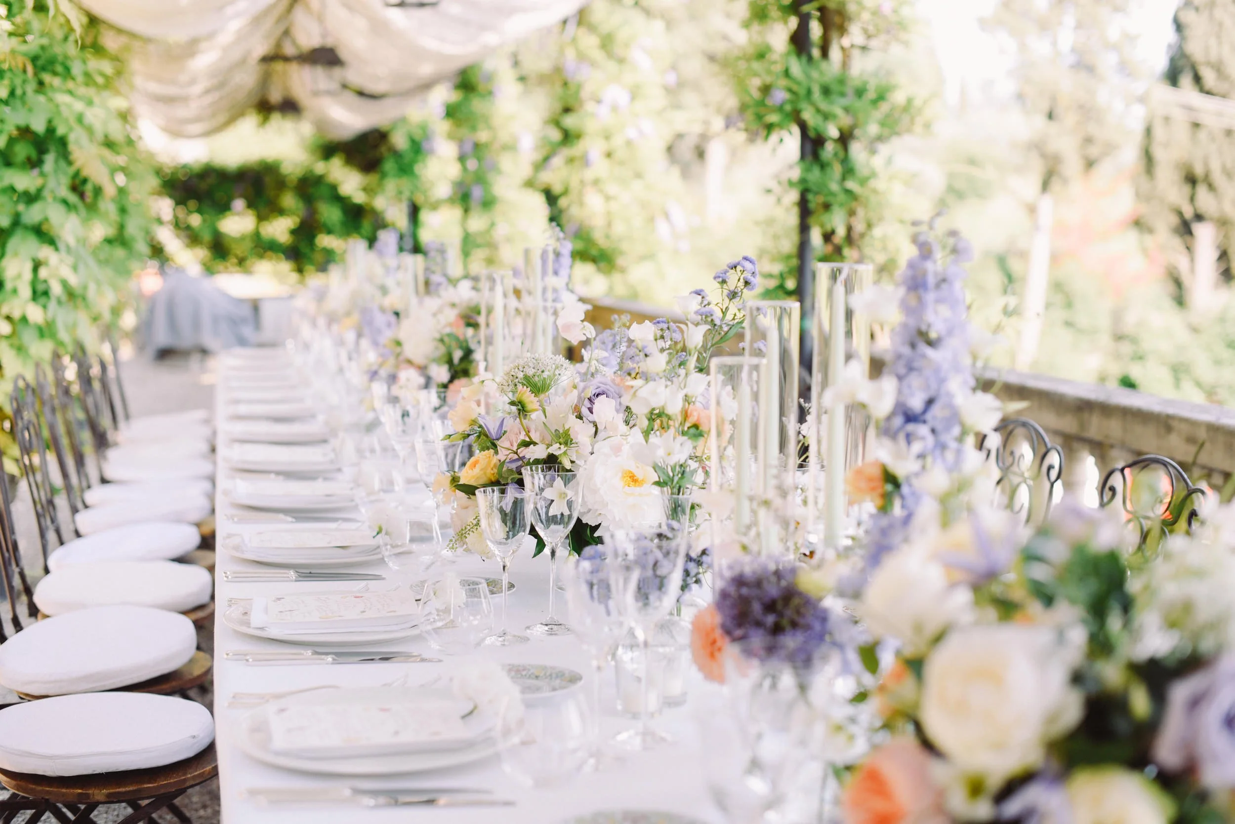 Long outdoor dining table decorated with floral centerpieces, surrounded by chairs with white cushions, set with glassware and plates, in a garden or courtyard setting with trees and greenery in the background.