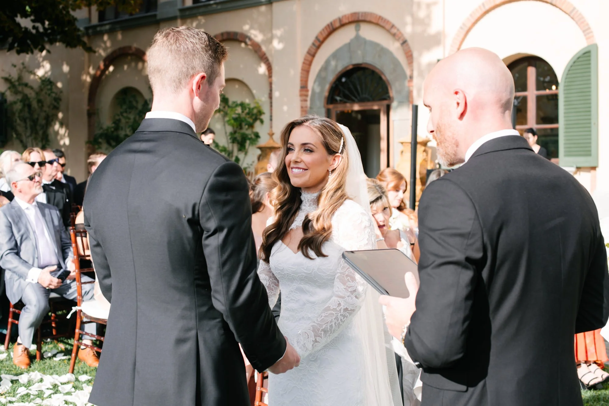 A bride and groom exchanging vows outdoors during a wedding ceremony, with a person officiating and seated guests in the background.