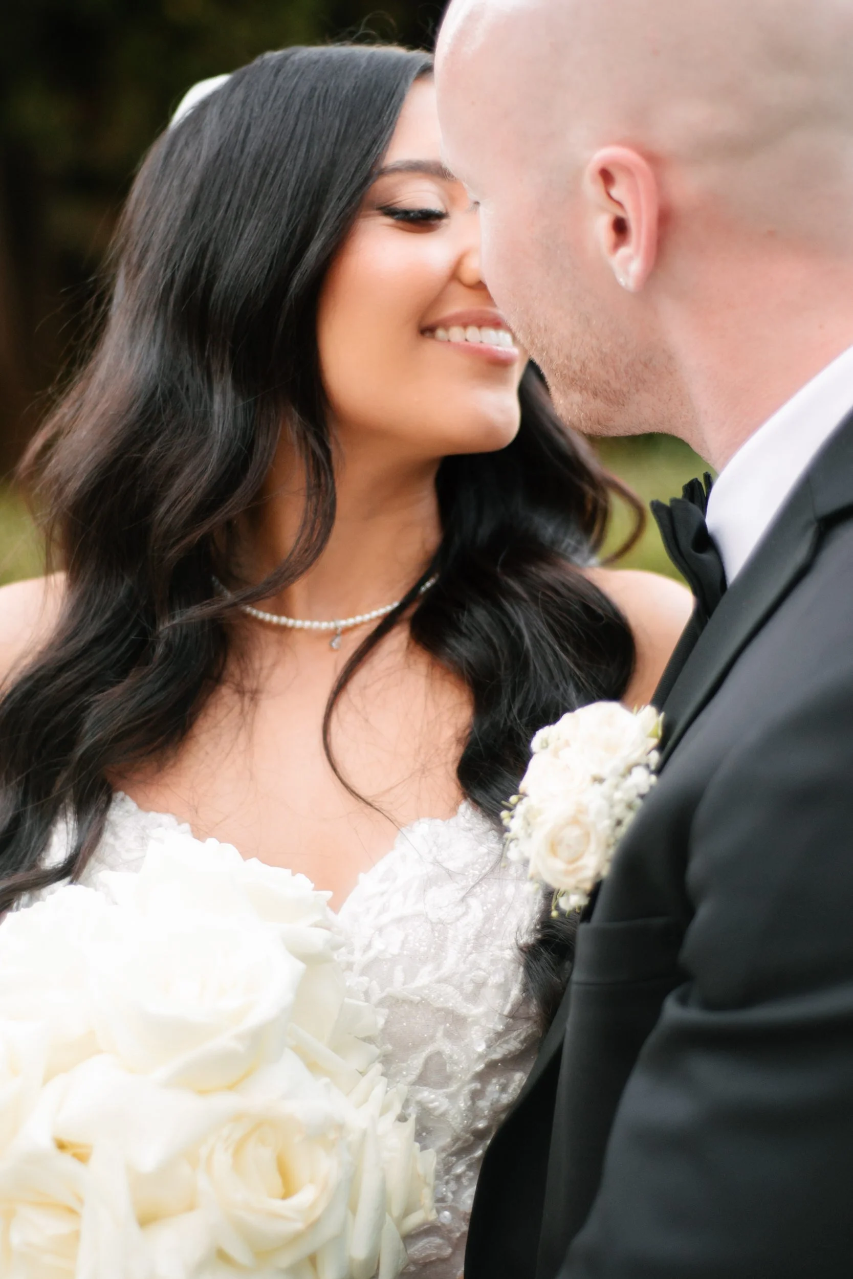 A bride and groom face each other closely on their wedding day, with the bride smiling and the groom's face partially visible, outdoors.