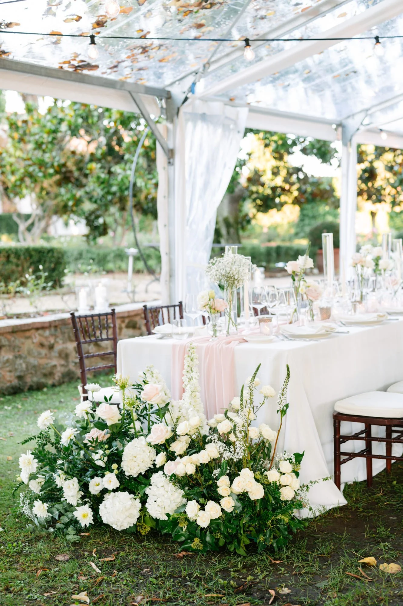 Elegant outdoor wedding reception setup with a long table decorated with white and blush flowers, candles, and glassware under a clear tent with string lights, surrounded by greenery and trees.