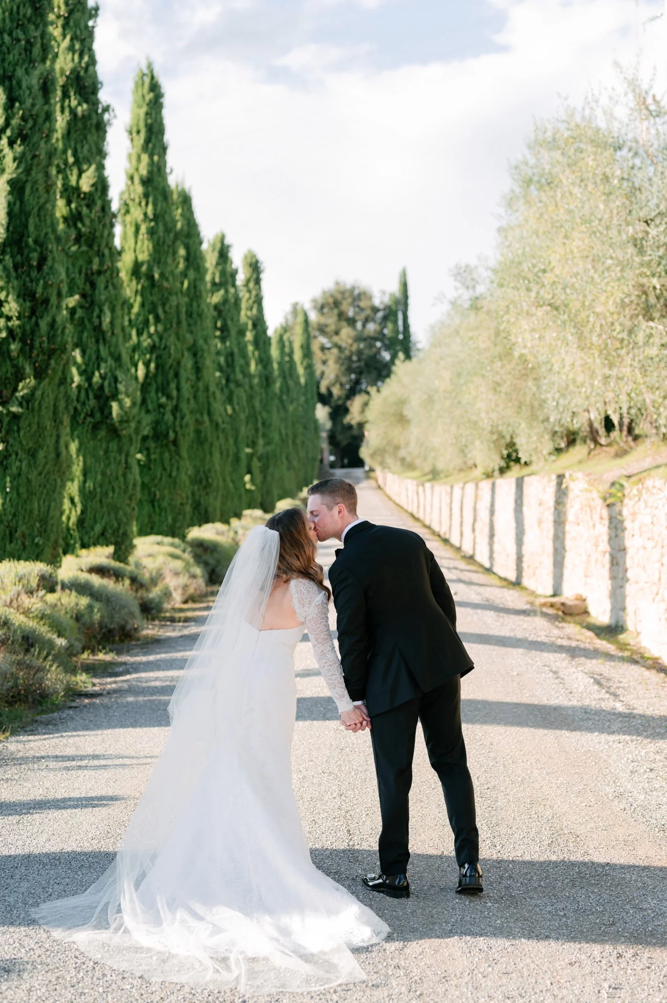 A bride and groom kiss and hold hands on a tree-lined road on a sunny day.