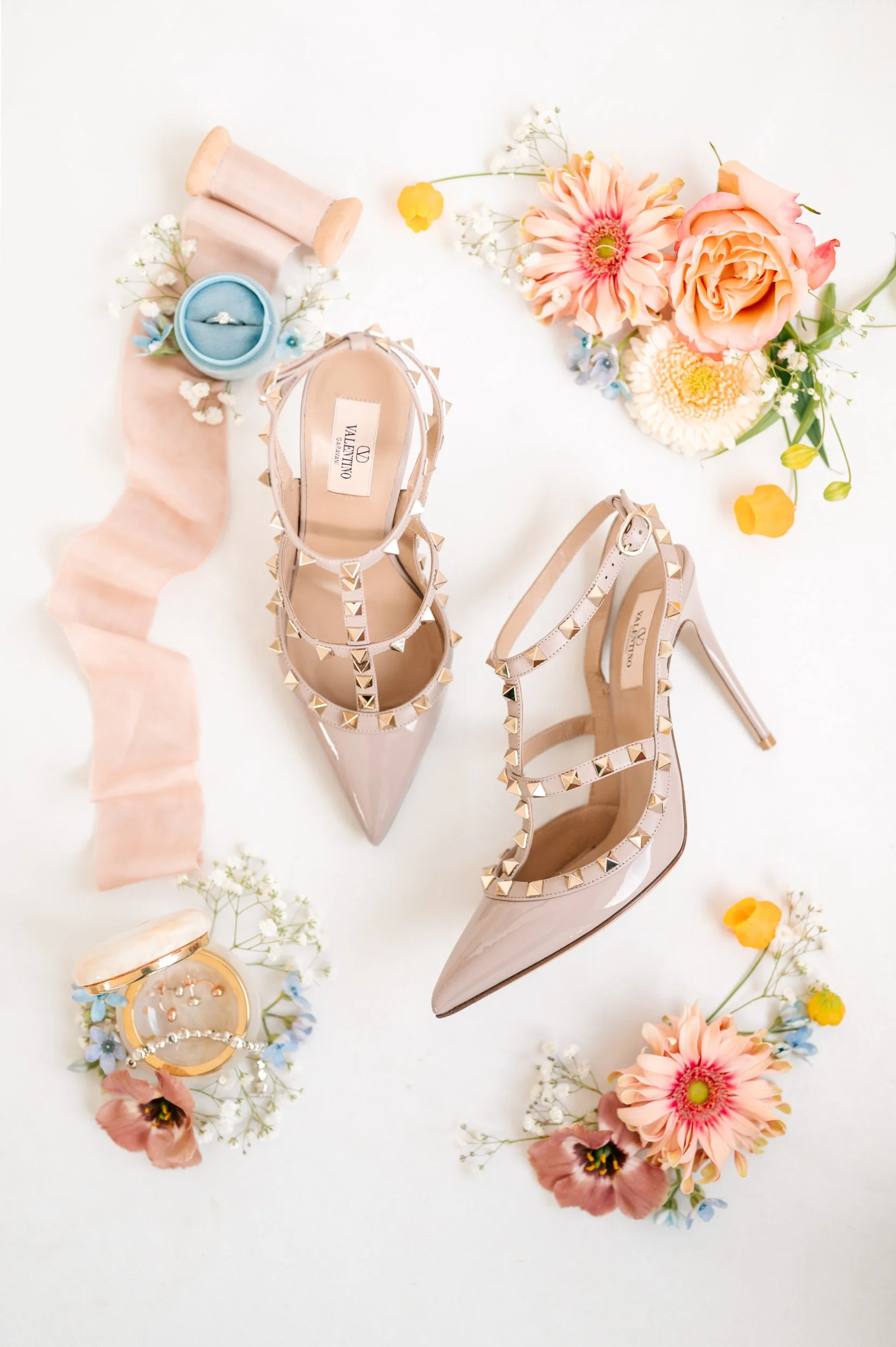 Pair of beige Valentino high-heeled shoes with gold pyramid studs, surrounded by colorful flowers, rings, and a pink ribbon.